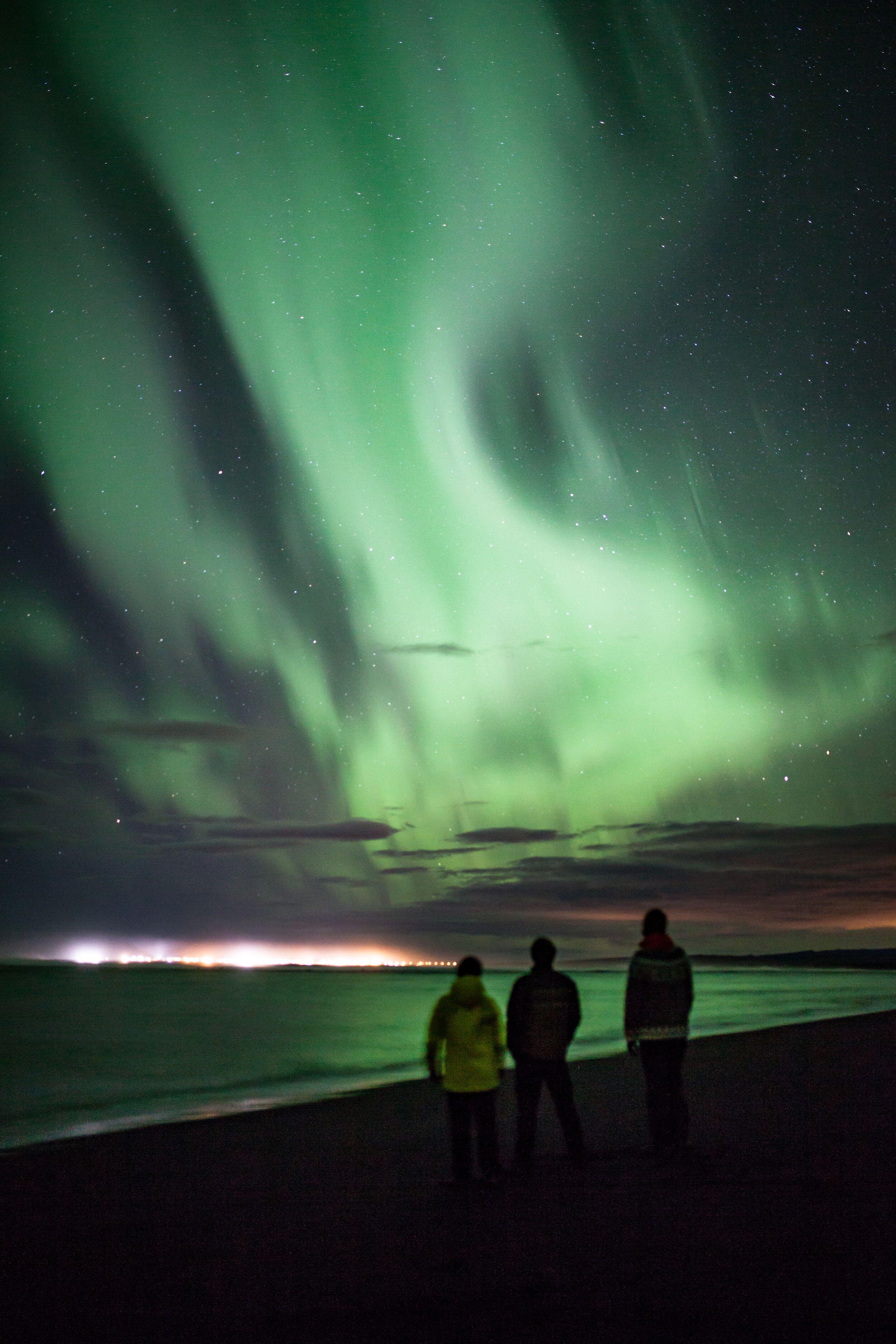 Three people standing on a dark shoreline, watching vibrant green Northern Lights illuminate the night sky.
