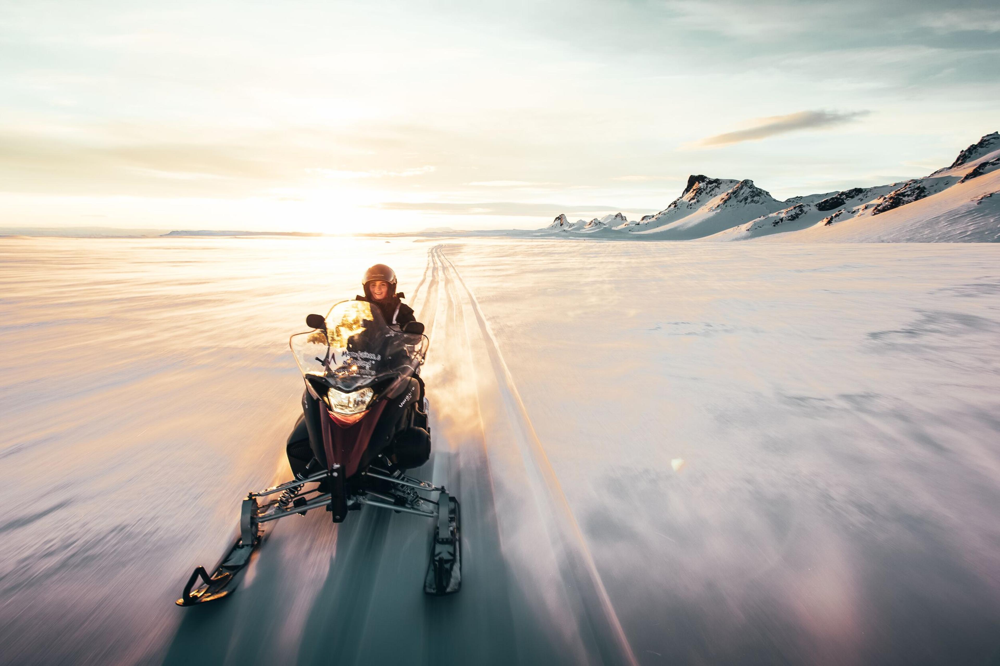 A person riding a snowmobile across a vast glacier, silhouetted against the radiant glow of a setting sun.