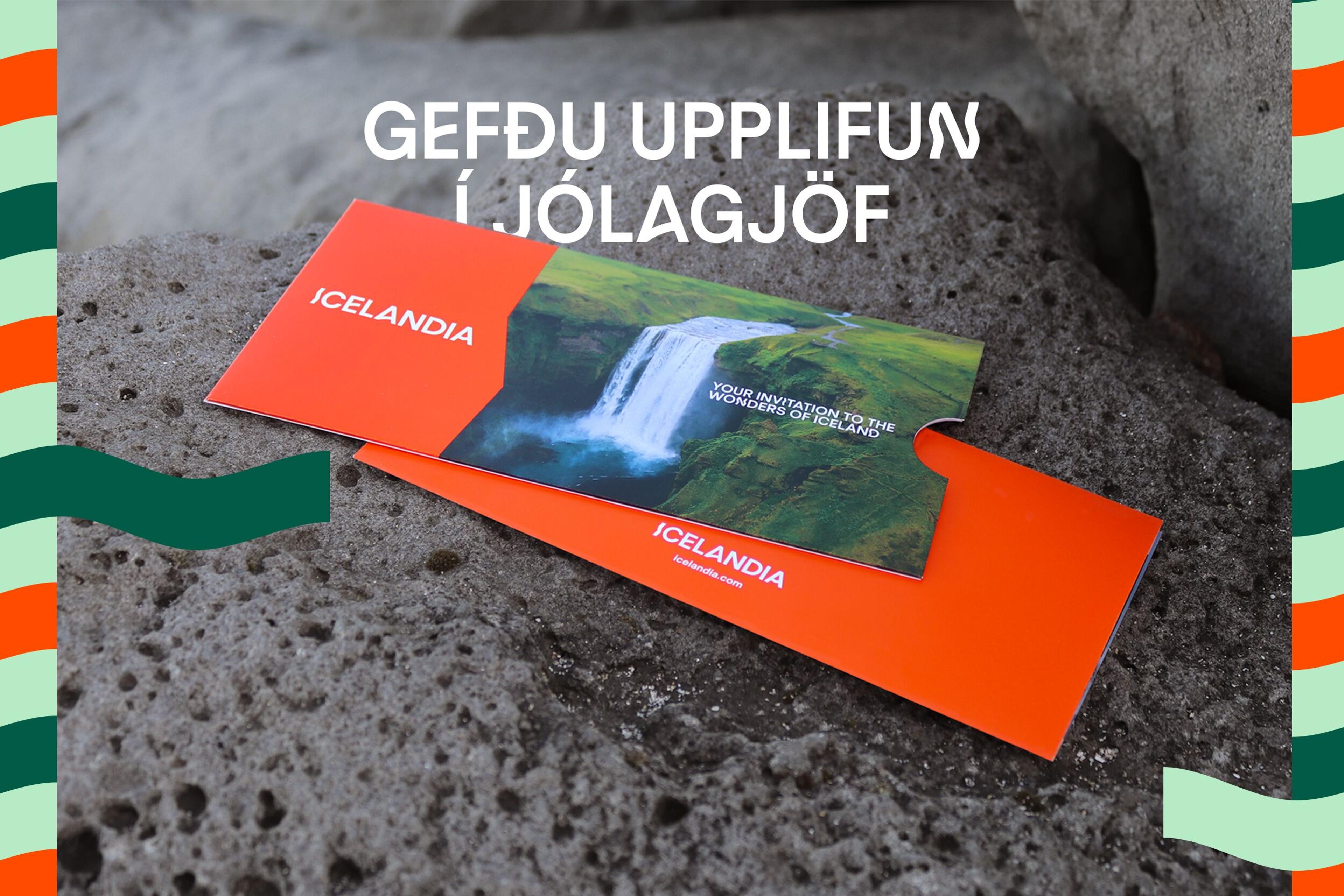 An orange Icelandia gift card with a waterfall image and "Your invitation to the wonders of Iceland" text, resting on rocks. Overlaid text in Icelandic reads "Give an experience as a Christmas gift."