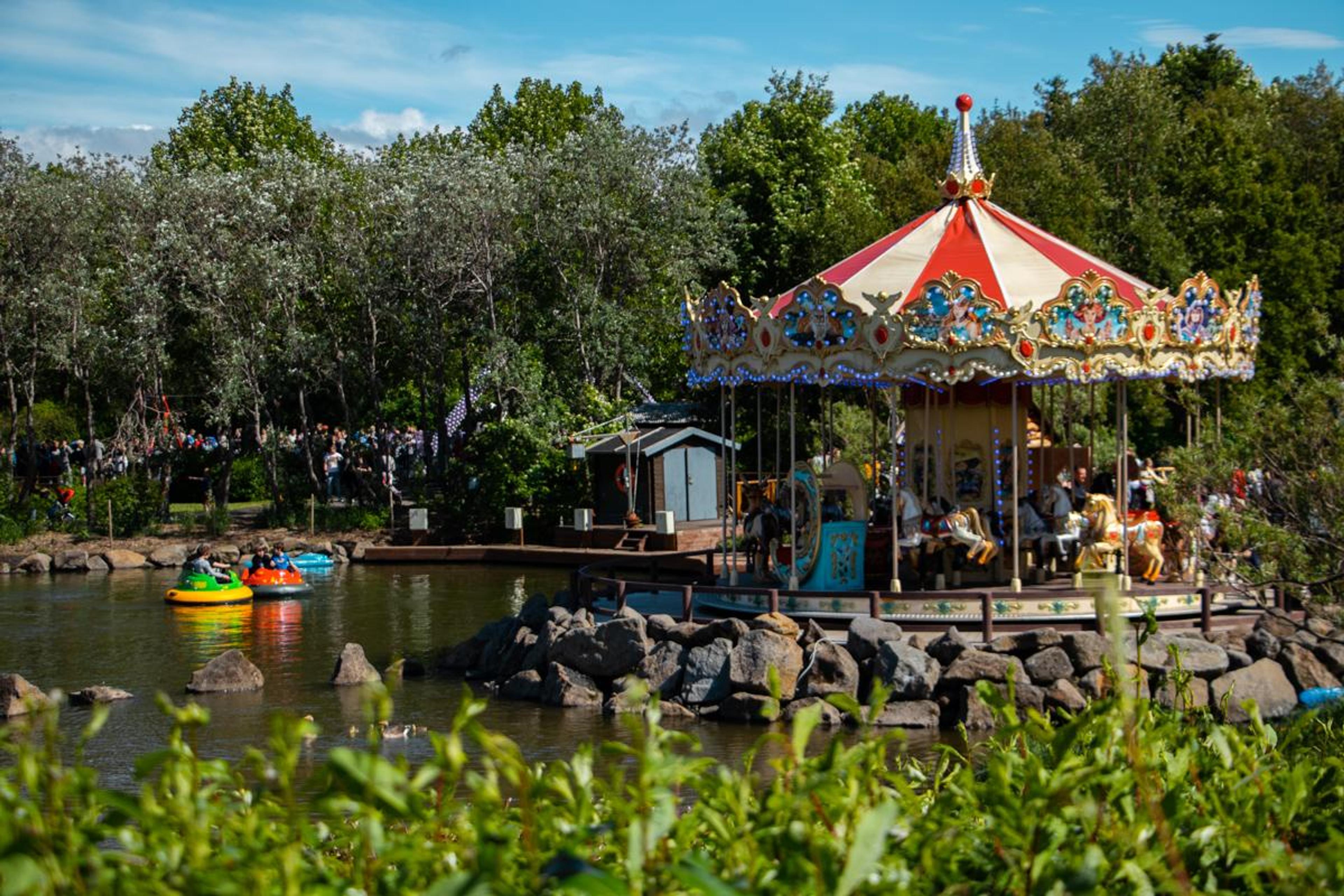 Colorful carousel by a pond in a lush park, with visitors enjoying a sunny day.