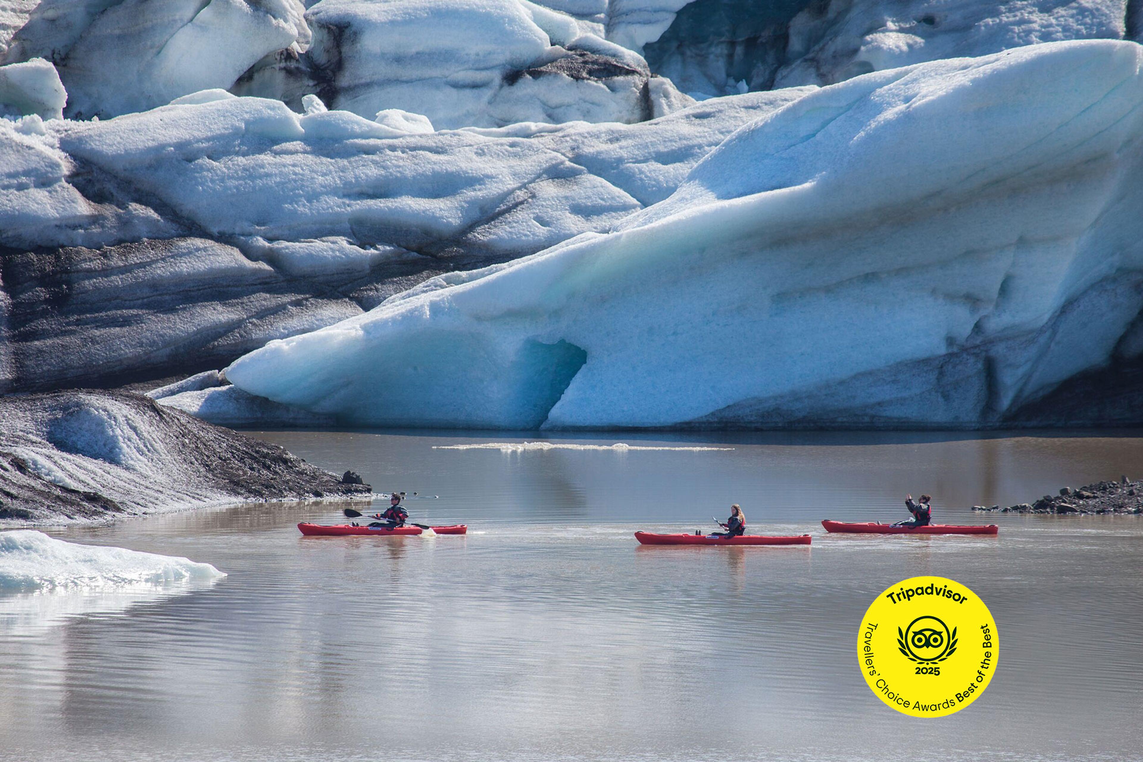 three people are kayaking in a lake in front of a large iceberg .