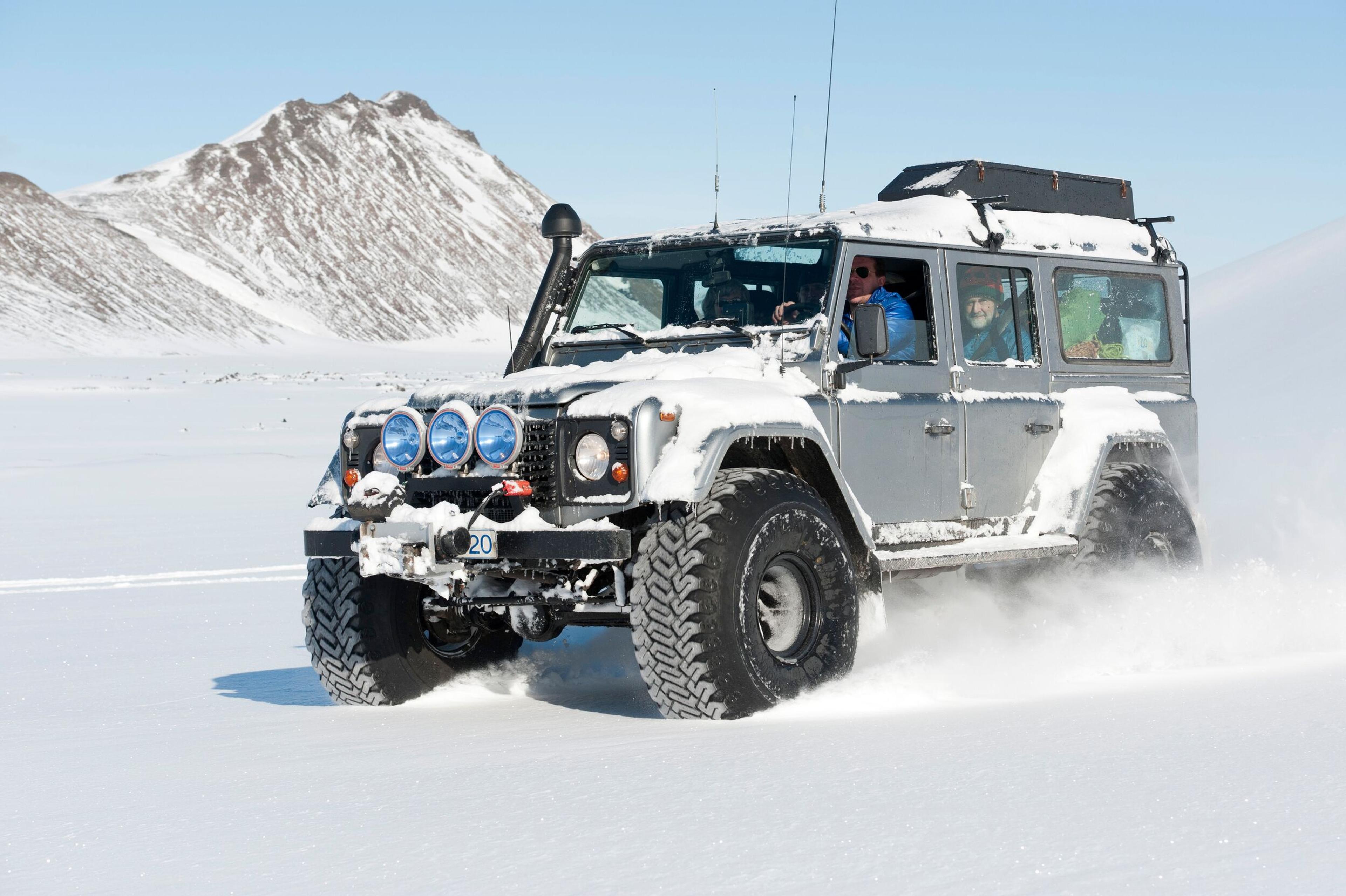 A large Jeep crossing in a snowy field against a backdrop of sunlit snowy mountains.