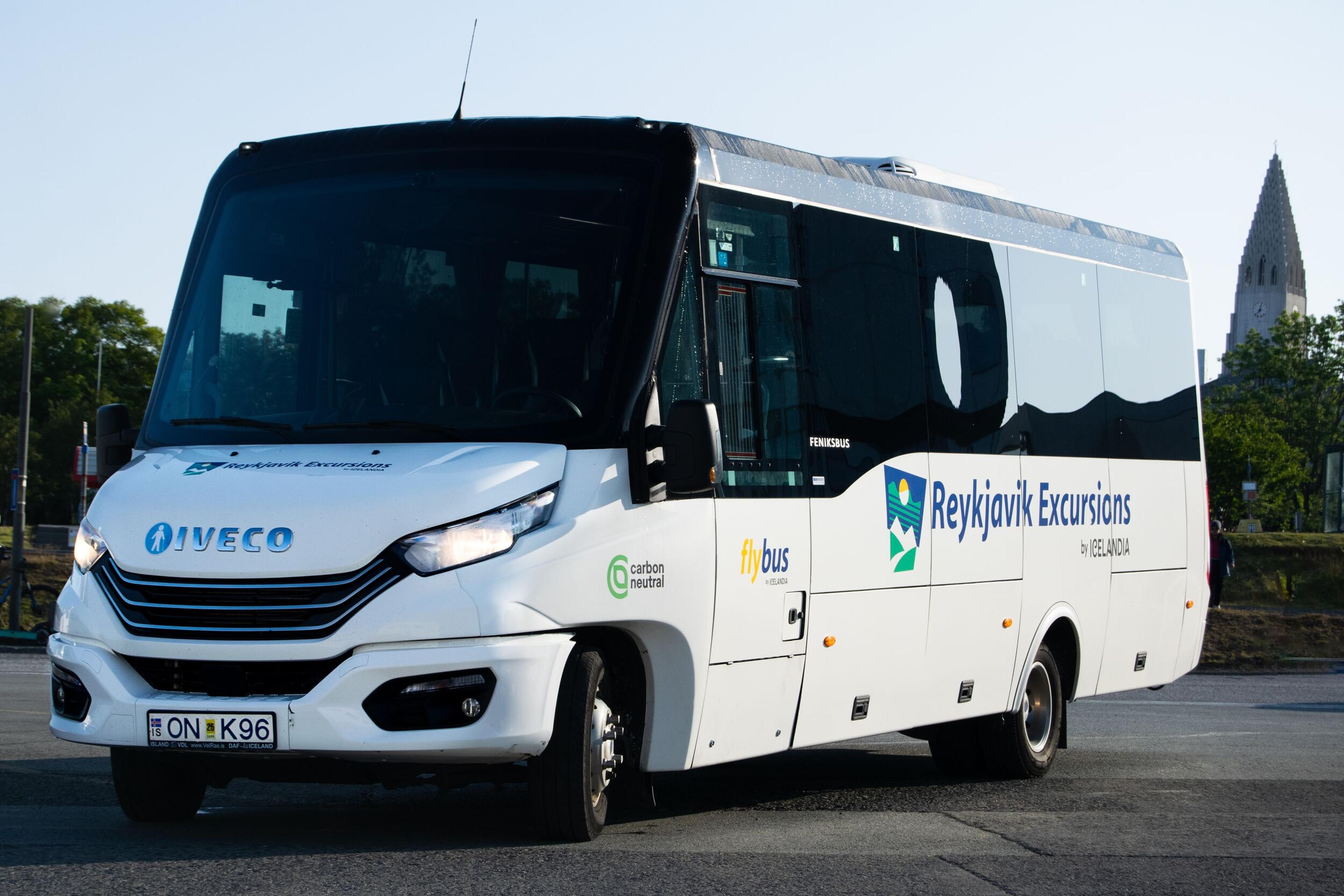A white Iveco Reykjavik Excursions Flybus with "carbon neutral" branding, with the Hallgrímskirkja church steeple in the background.