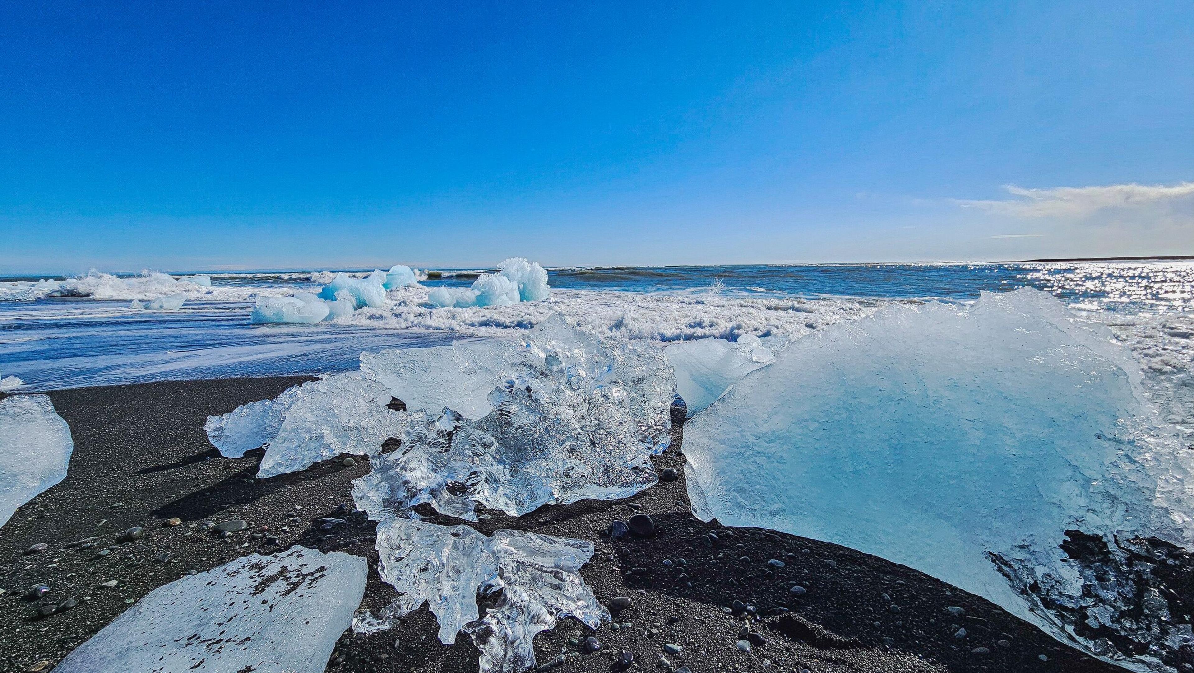 Clear and blue ice chunks scattered on a black sand beach with ocean waves under a bright blue sky.