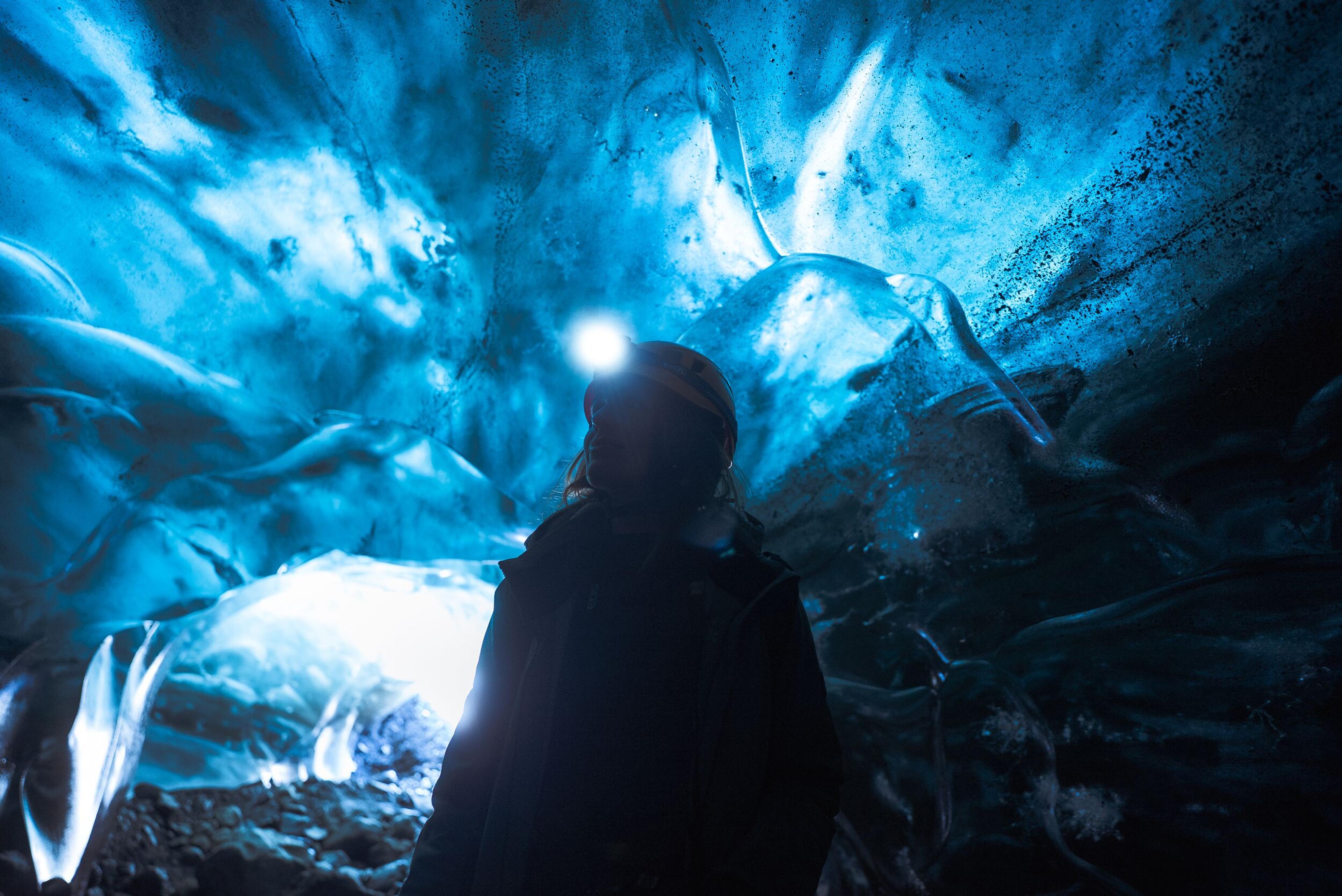 Person with a headlamp inside a glowing blue ice cave.