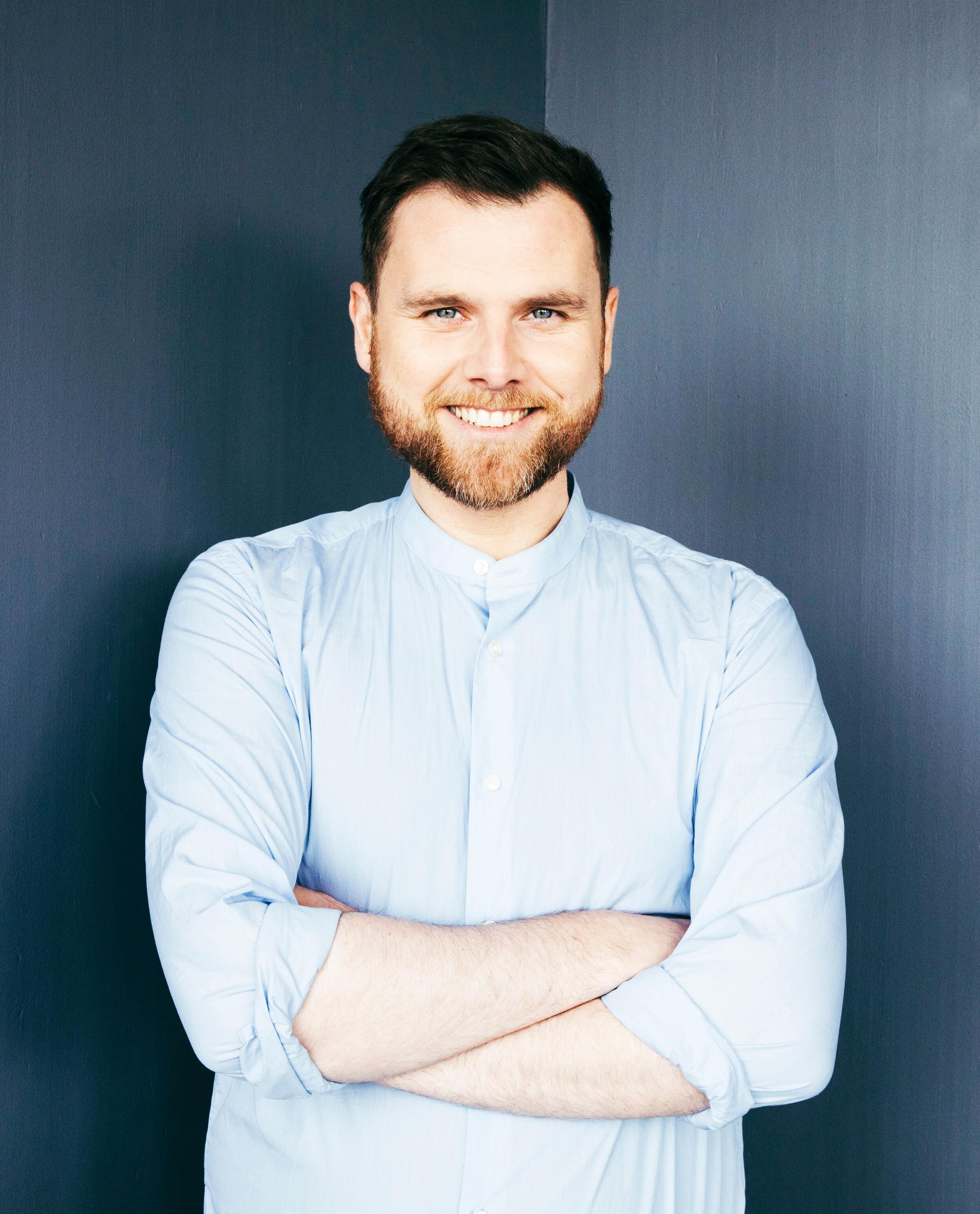 A man with short brown hair and a beard, wearing a light blue button-up shirt, smiles with his arms crossed.