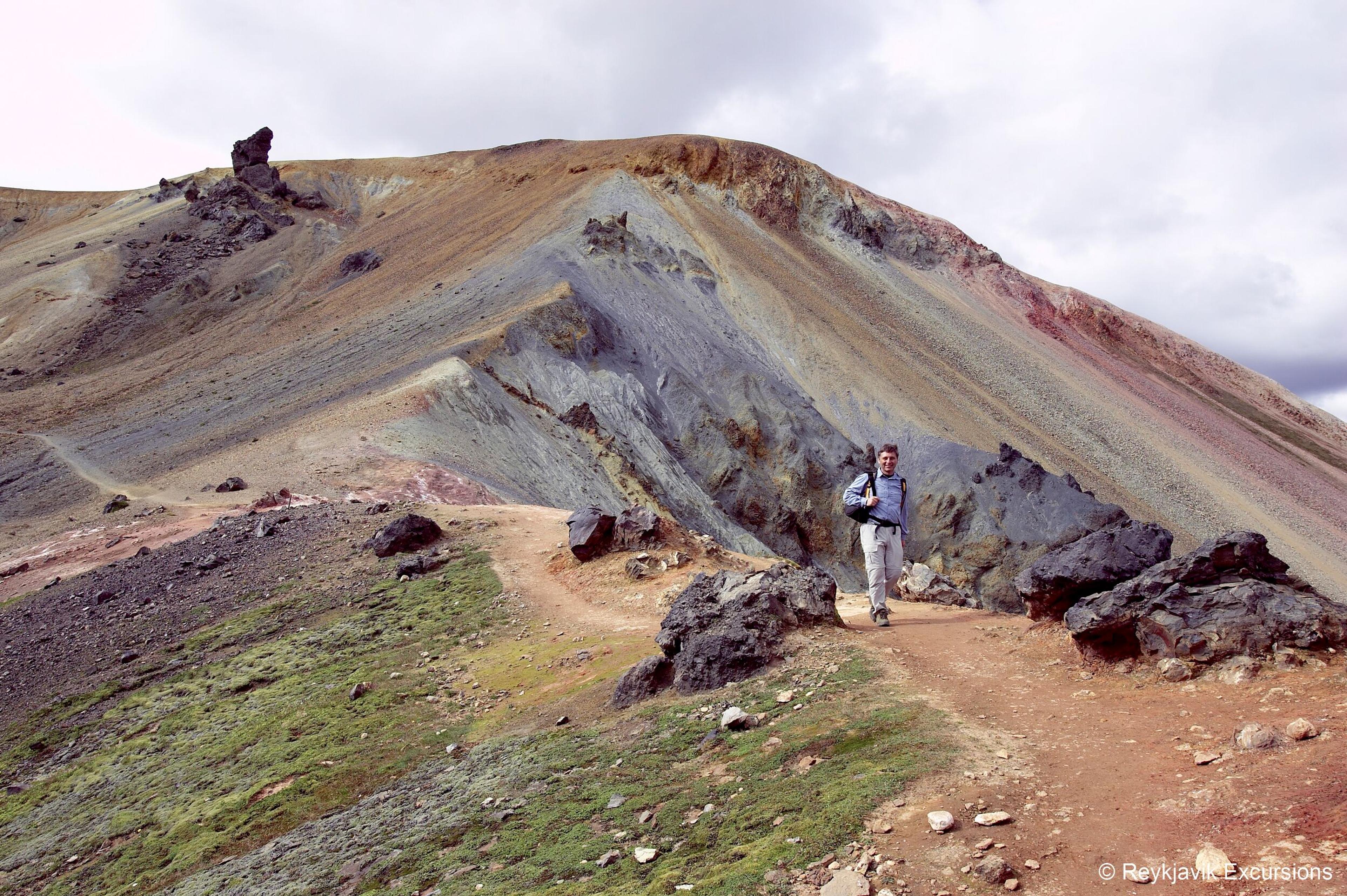 A hiker walks on a dirt path through the colorful, volcanic mountains of Brennisteinsalda, Landmannalaugar.