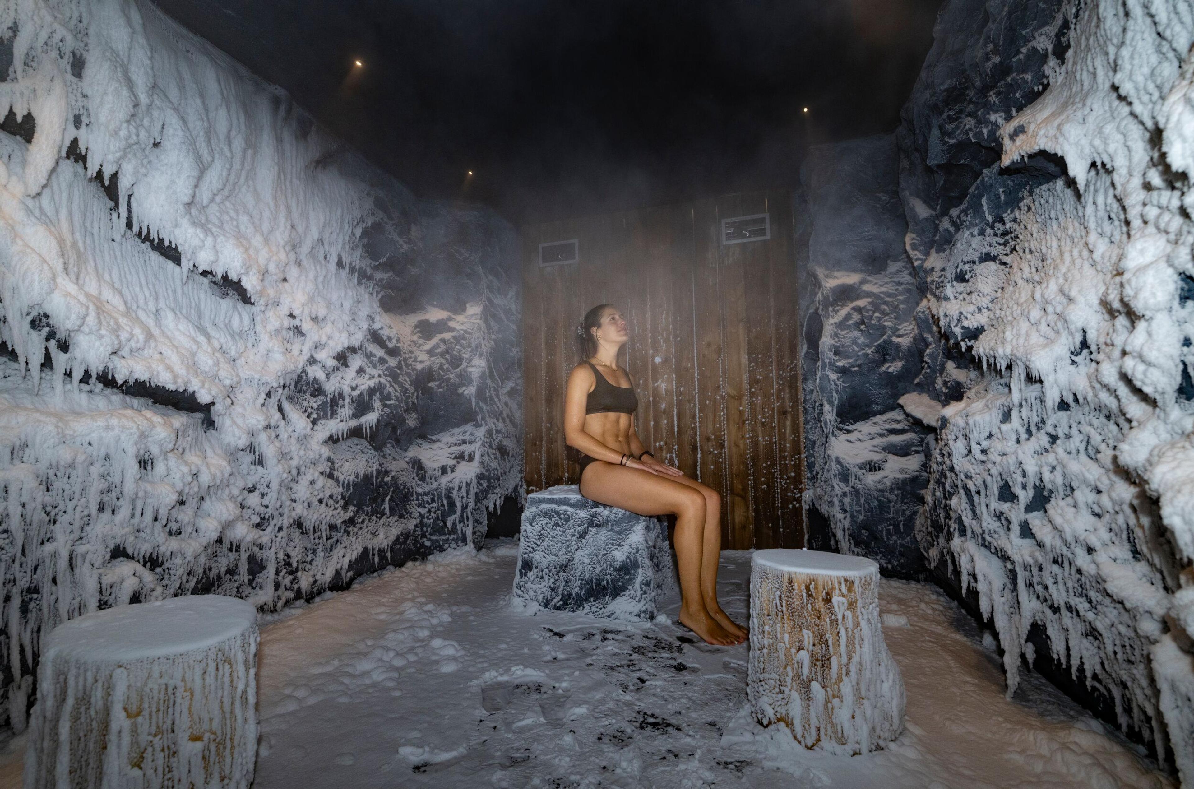 A woman in a bikini sitting on an ice block in a misty room with frozen, icicle-covered walls.