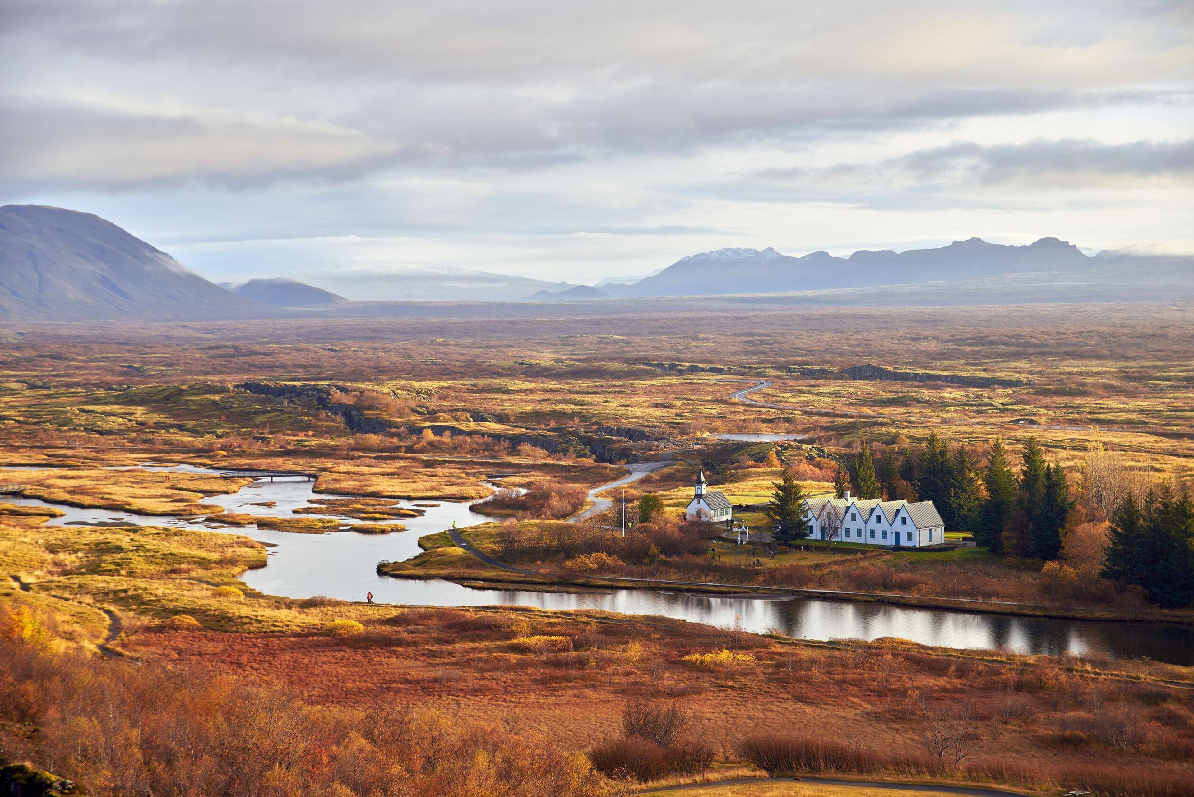 Scenic view of Thingvellir National Park in Iceland, featuring autumn colors, winding rivers, and a small cluster of buildings amidst a vast landscape.