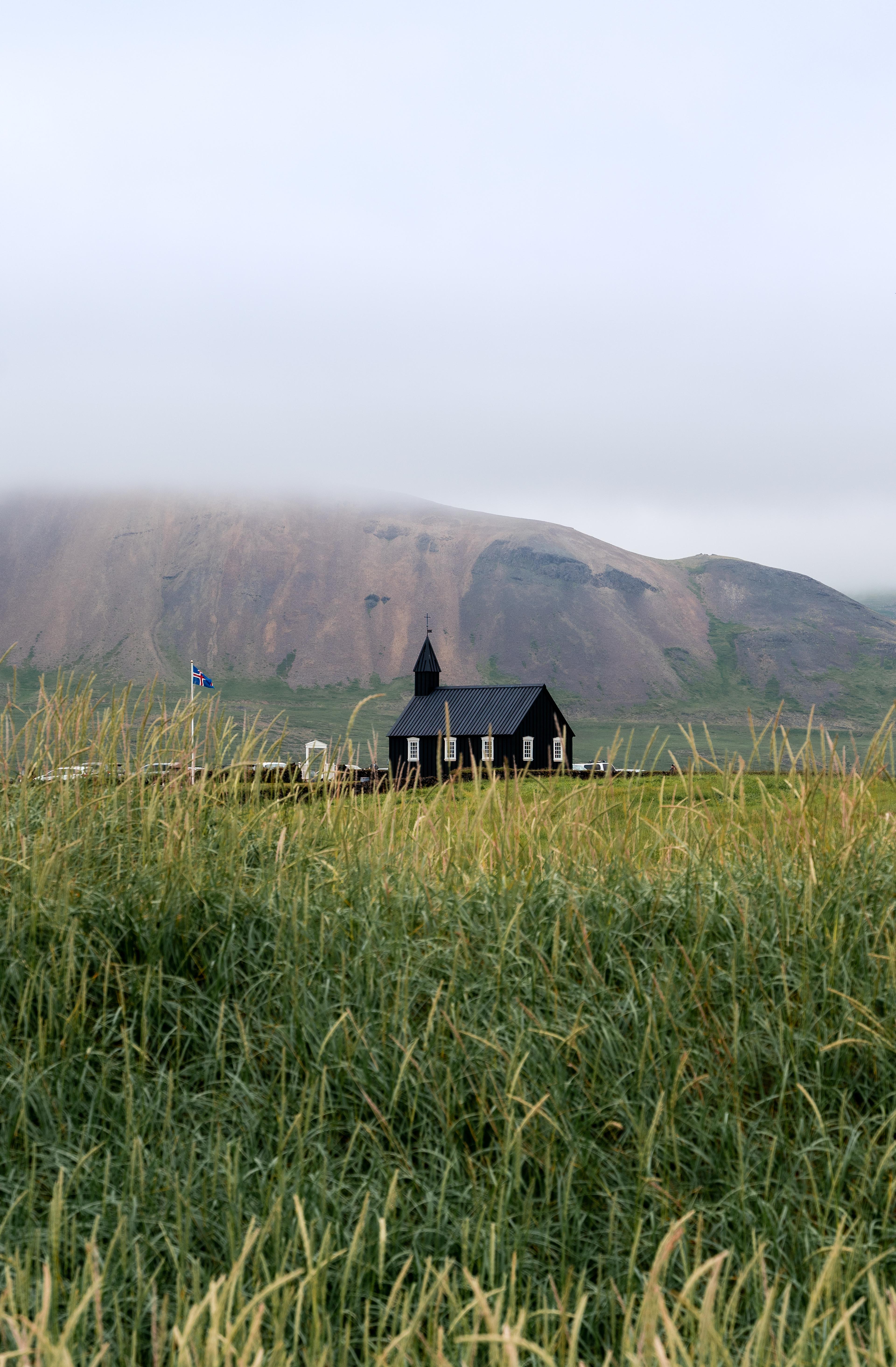 A black church with white windows stands in a green field with tall grass, a misty mountain, and an Icelandic flag behind it.