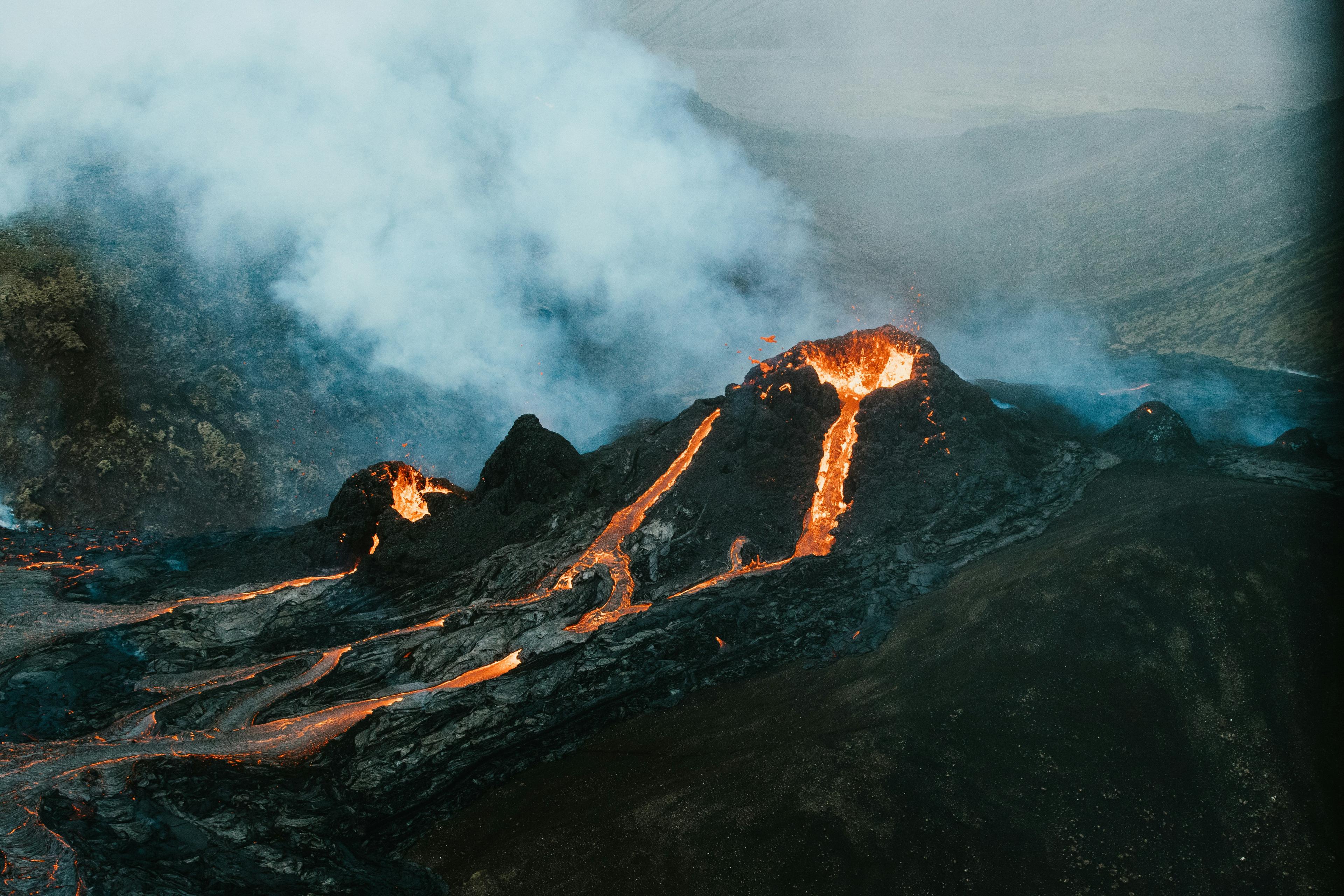 Aerial view of an active volcano with smoke and lava flows against a stormy sky.