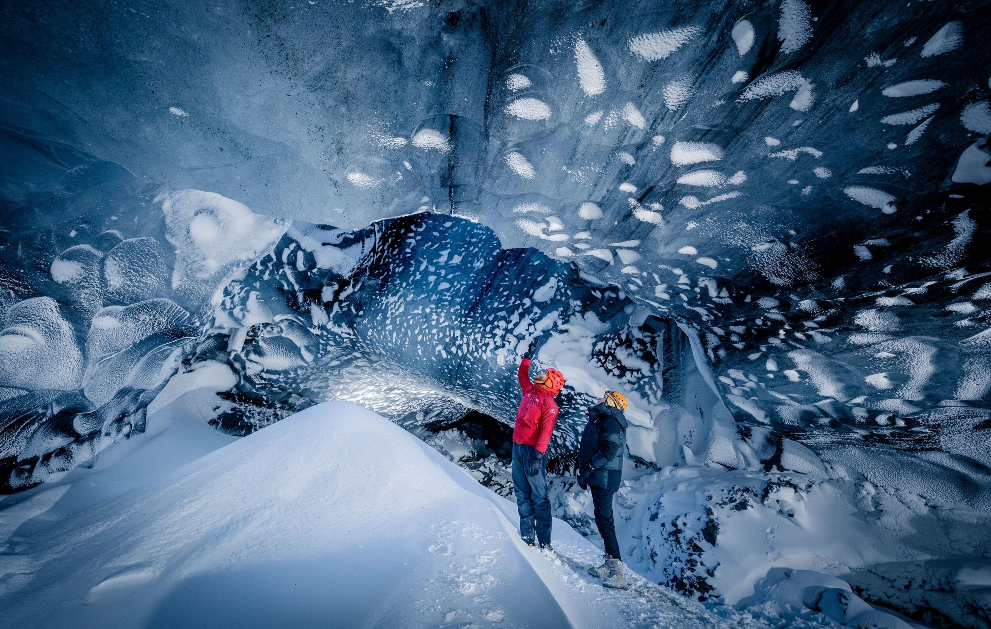 Two people in bright jackets standing at the entrance of a vast ice cave, with one person pointing towards the inside. The cave's inner walls glisten with layers of ice, and the snow on the ground enhances the cave's ethereal beauty.