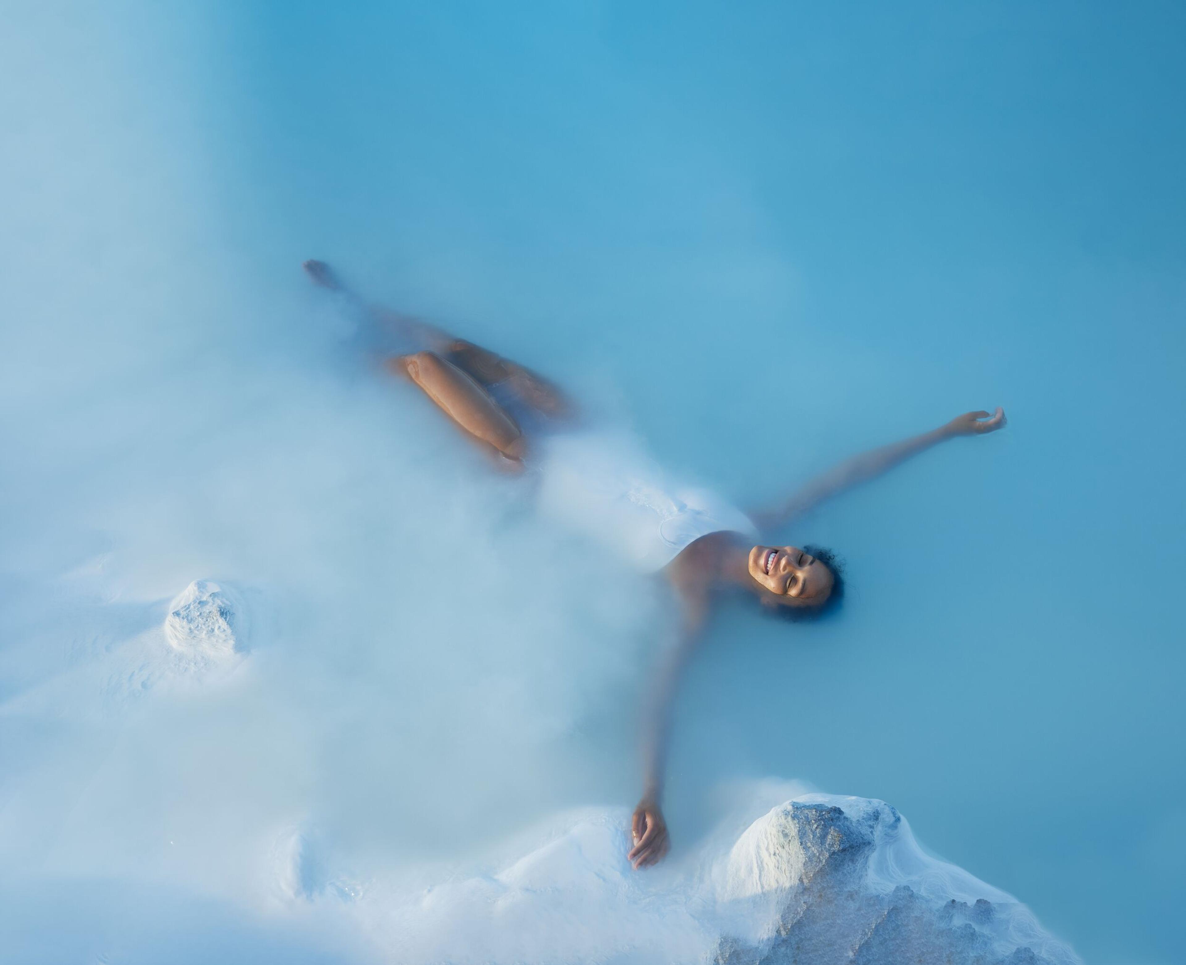 Woman relaxing and floating in the milky blue geothermal waters of Iceland's Blue Lagoon, surrounded by mist and white rocks.