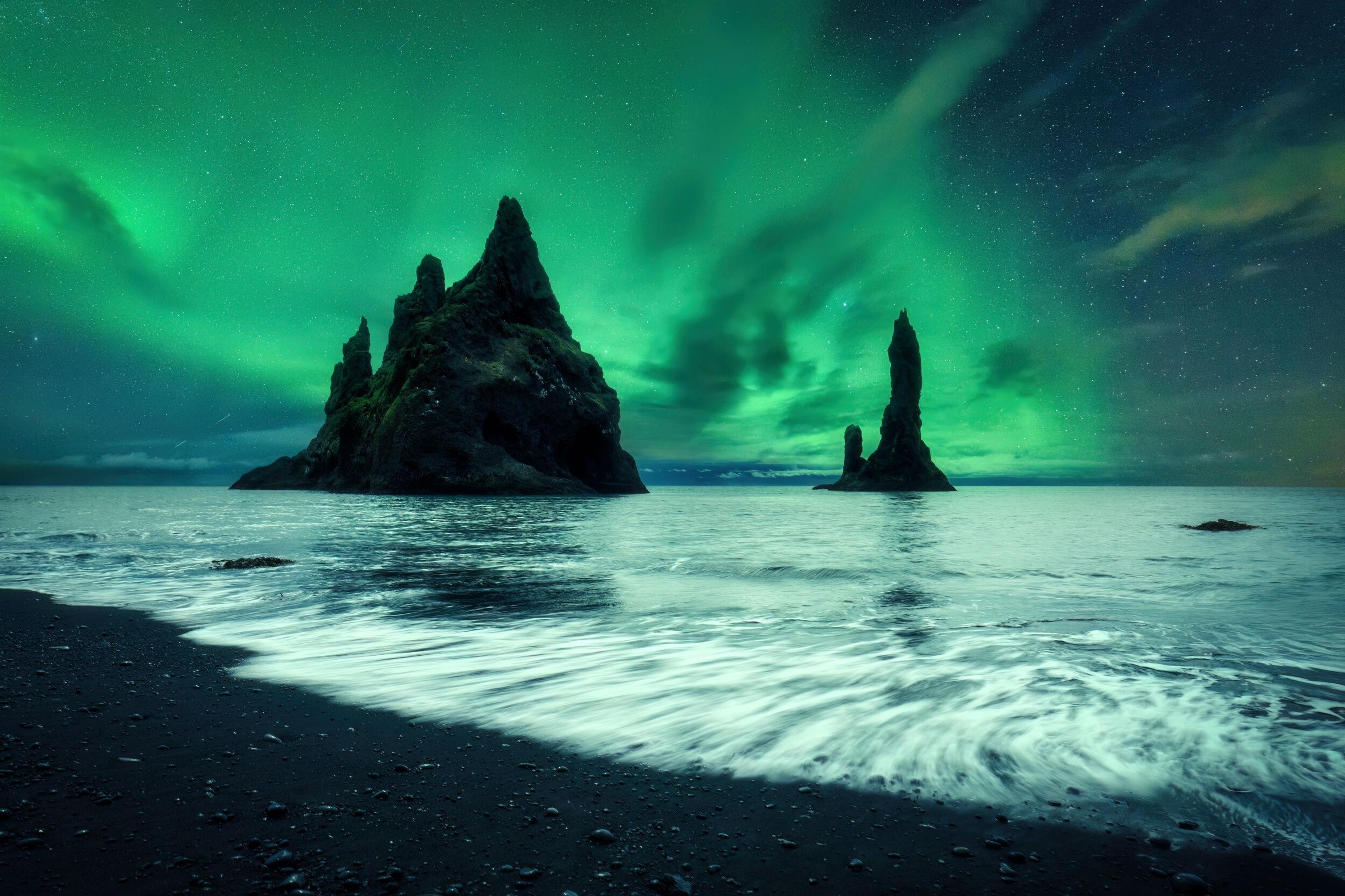 Green aurora borealis over a black sand beach with sea stacks in the ocean.