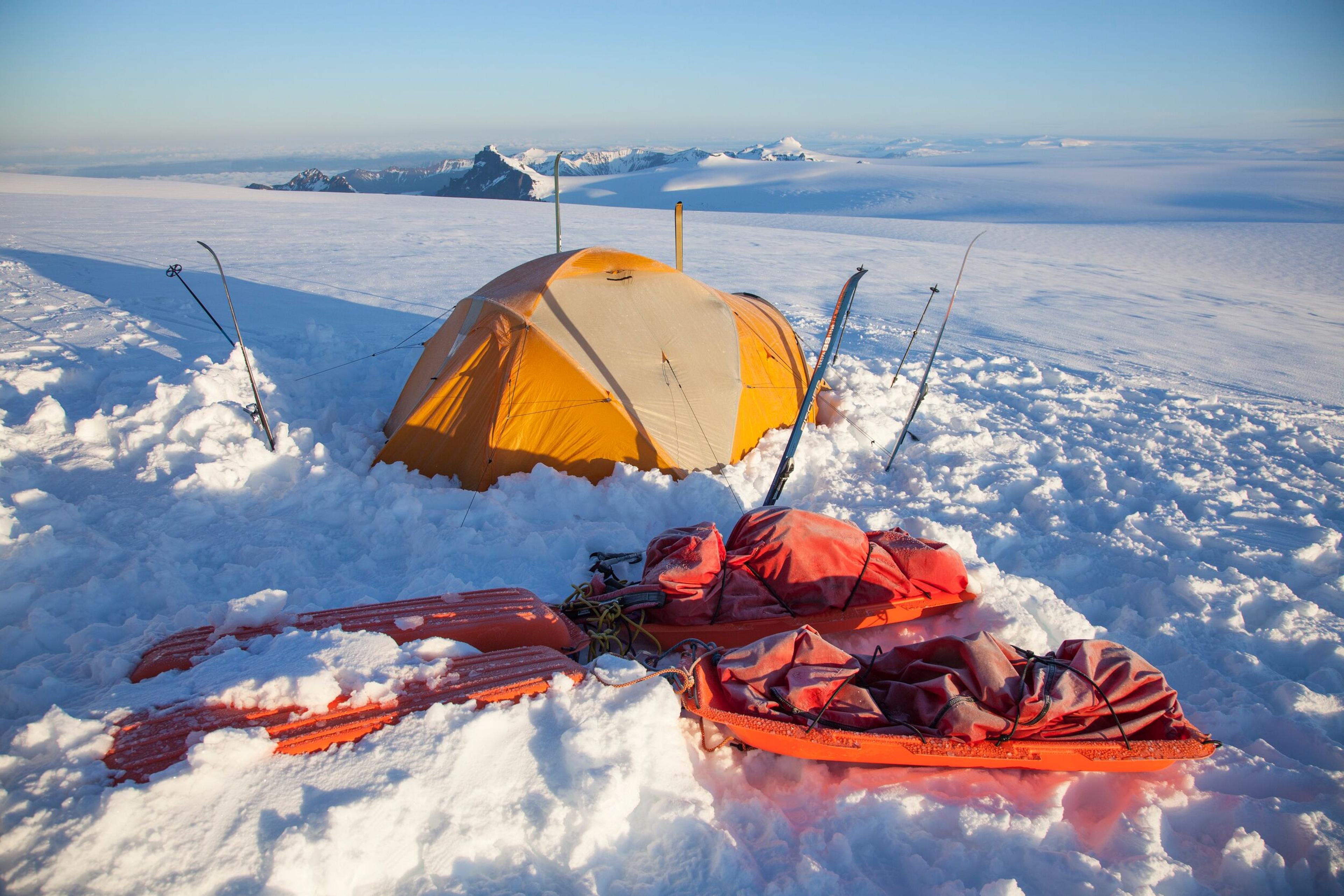 : A bright yellow tent stands pitched in a vast snowfield on a clear, sunny day, with skiing equipment in the foreground.