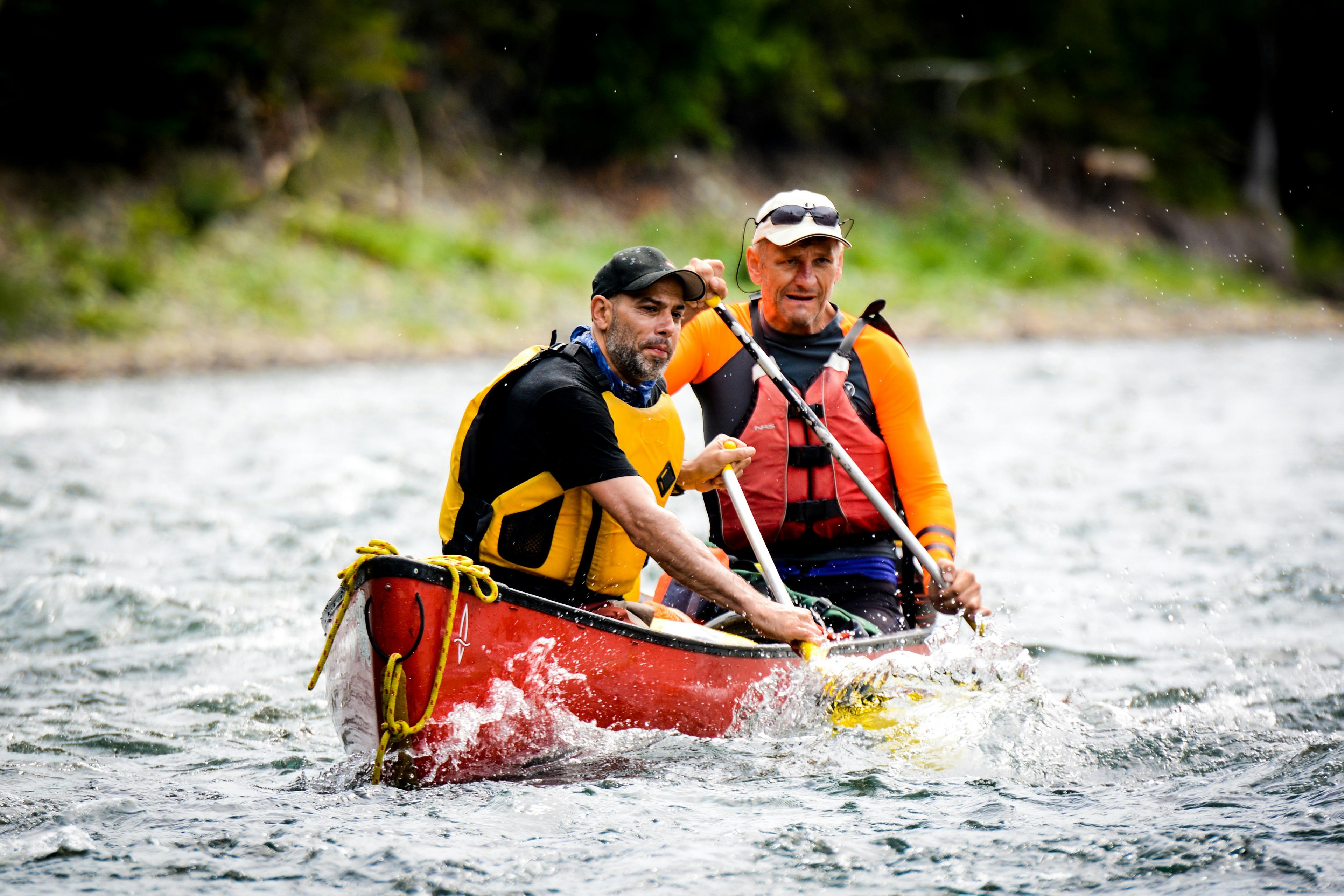 Two men paddle a red canoe on choppy water, wearing life vests and determined expressions, ideal for kayaking in Iceland.