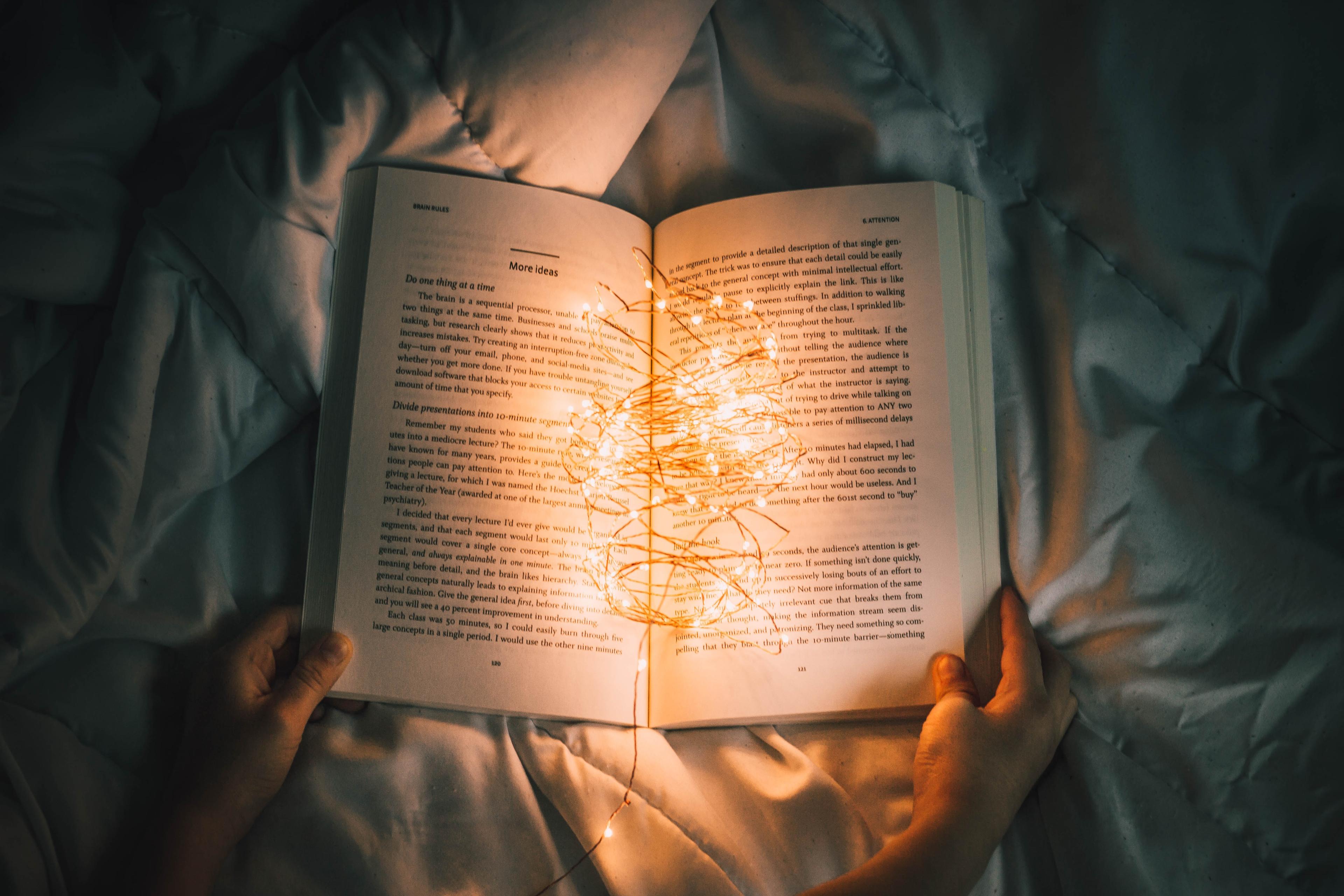 Hands holding an open book with a glowing string of fairy lights resting on the pages, atop a bed with grey sheets.