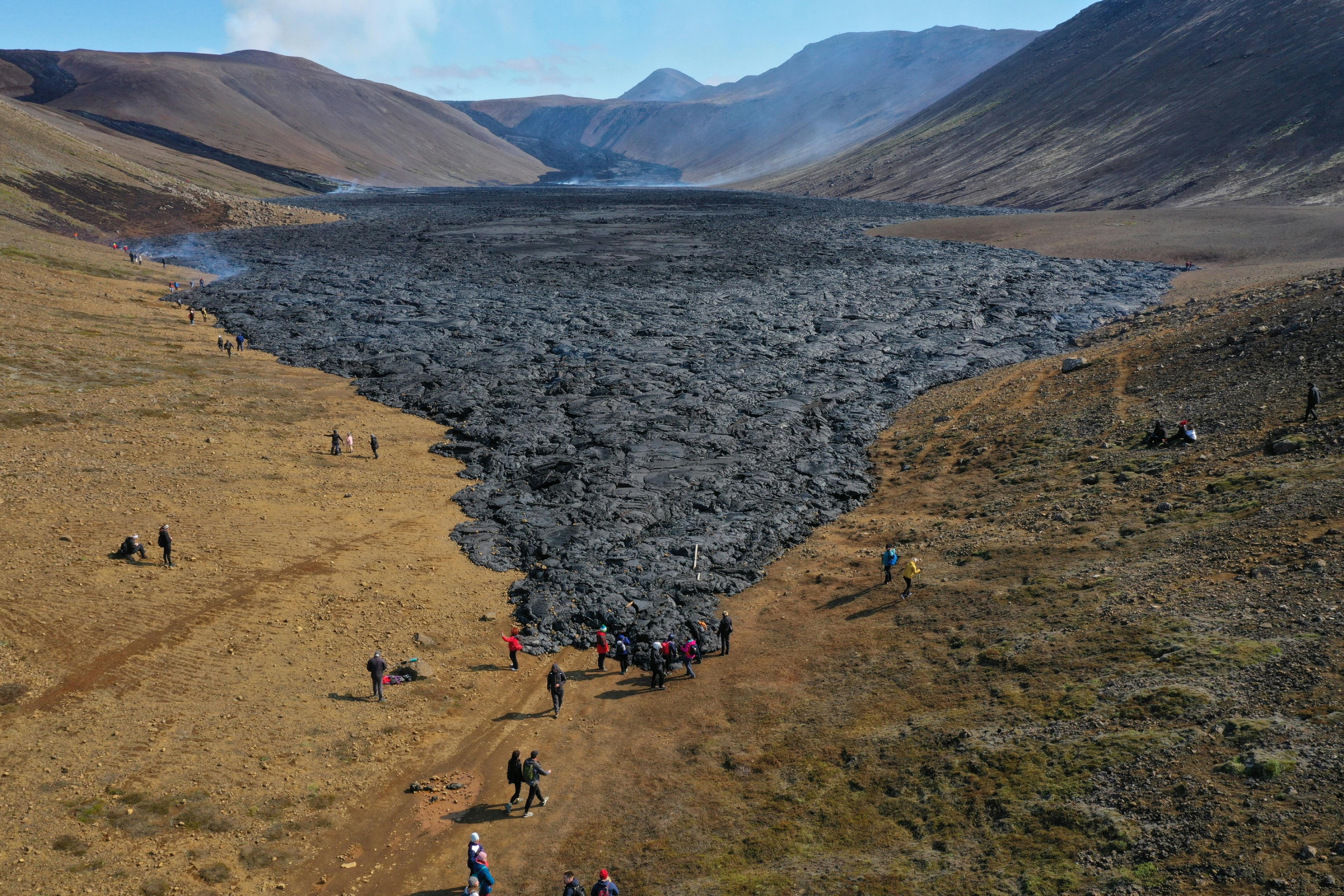 Aerial view of tourists exploring the edge of a dark lava field in a broad valley with mountains and clear skies in the background.