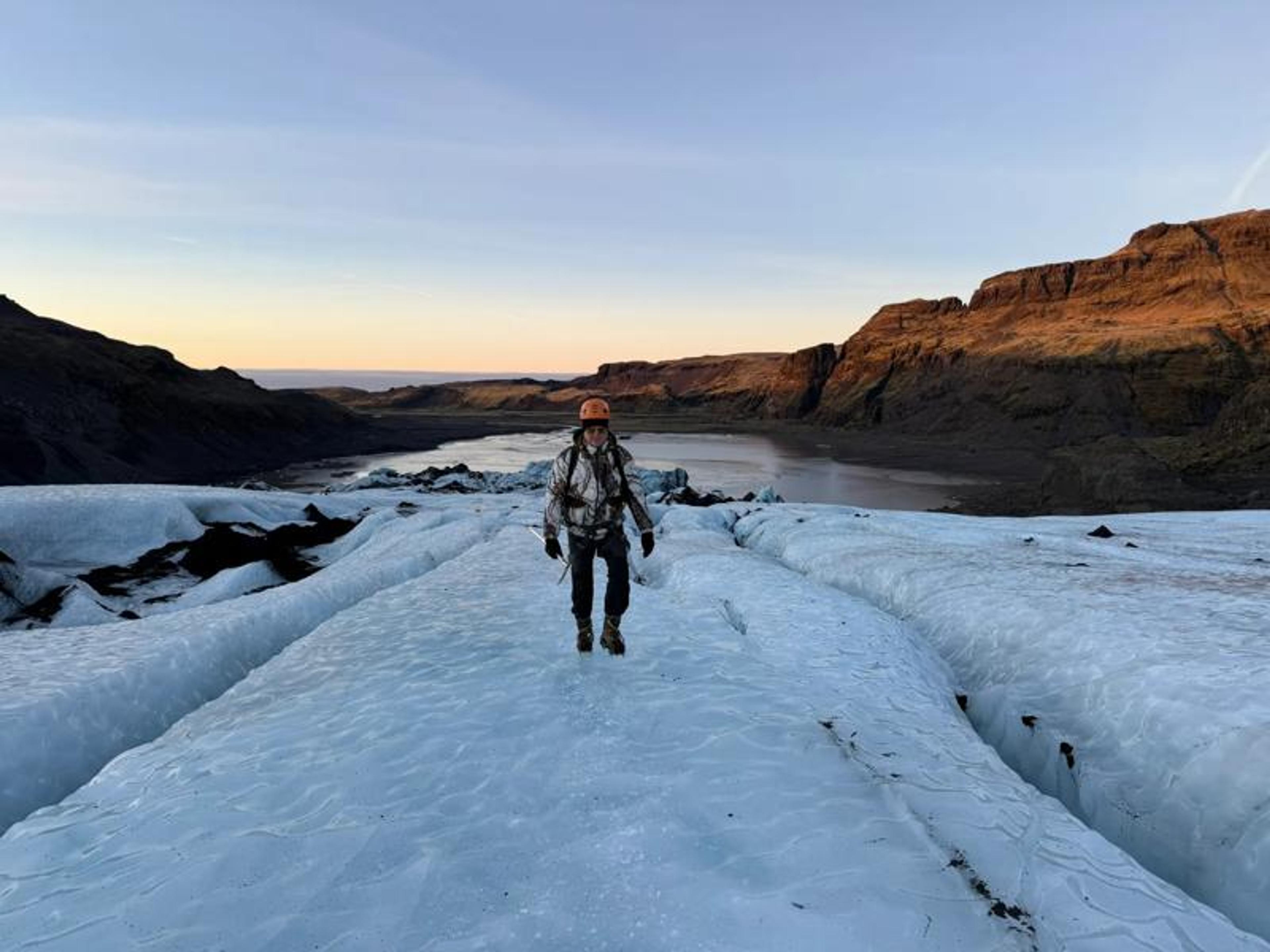 A glacier hiker wearing a helmet and carrying an ice axe walks on blue-white ice, with dark mountains and a lake under a warm sky.