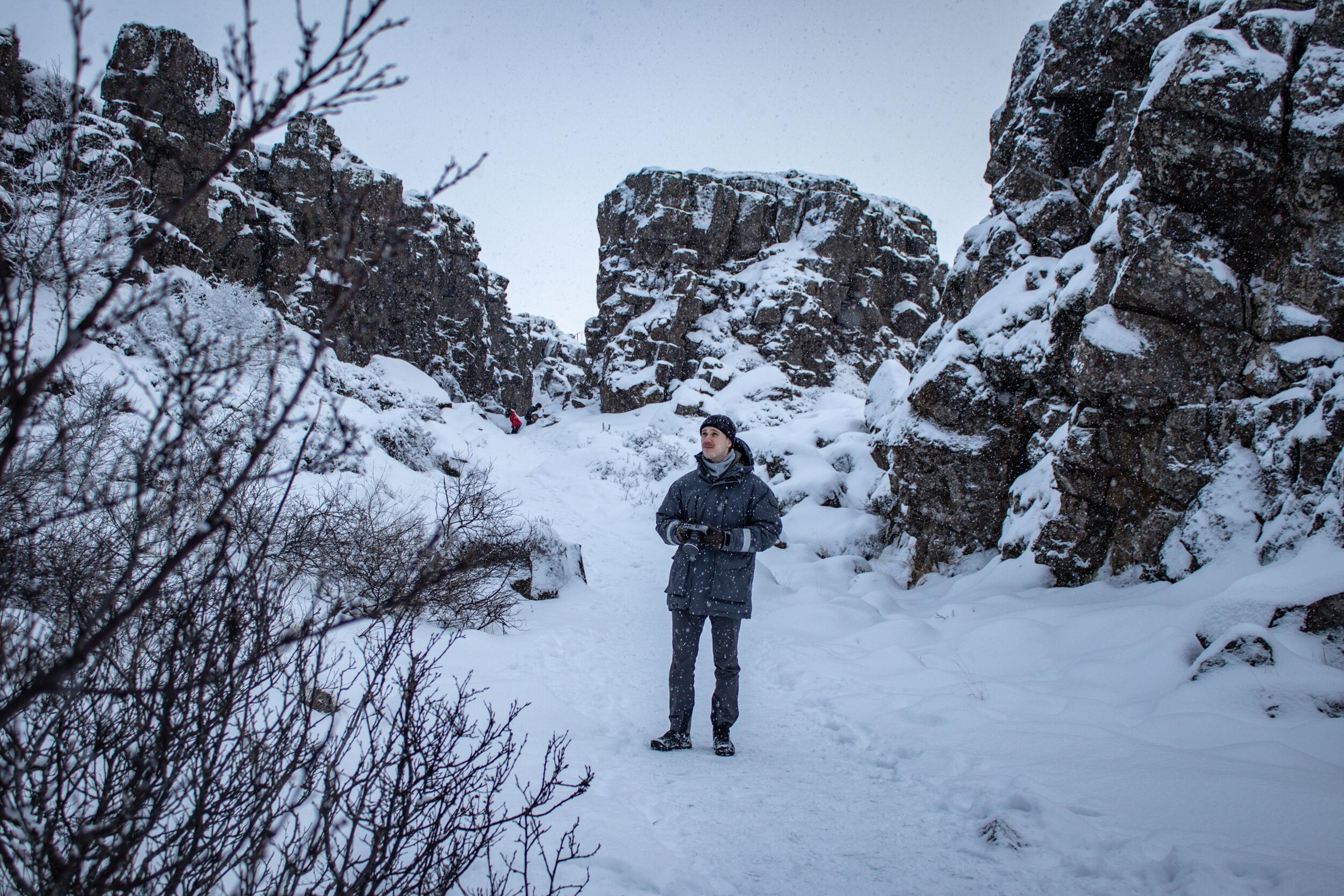 A man in winter clothes stands in a snowy gorge flanked by tall, snow-covered rock formations.
