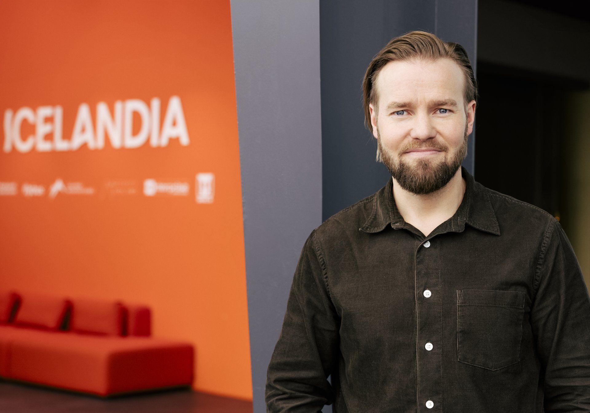 A man with light hair and a beard, wearing a dark brown shirt, smiles against a gray and orange background.