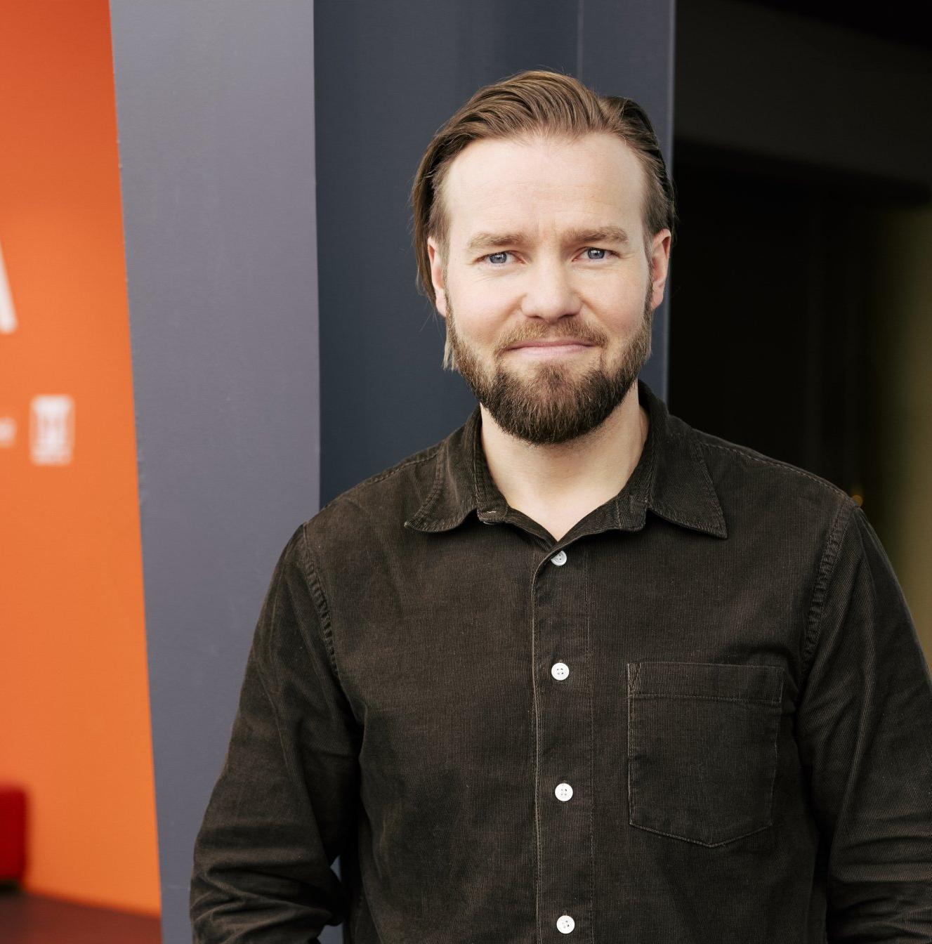 A man with light hair and a beard, wearing a dark brown shirt, smiles against a gray and orange background.