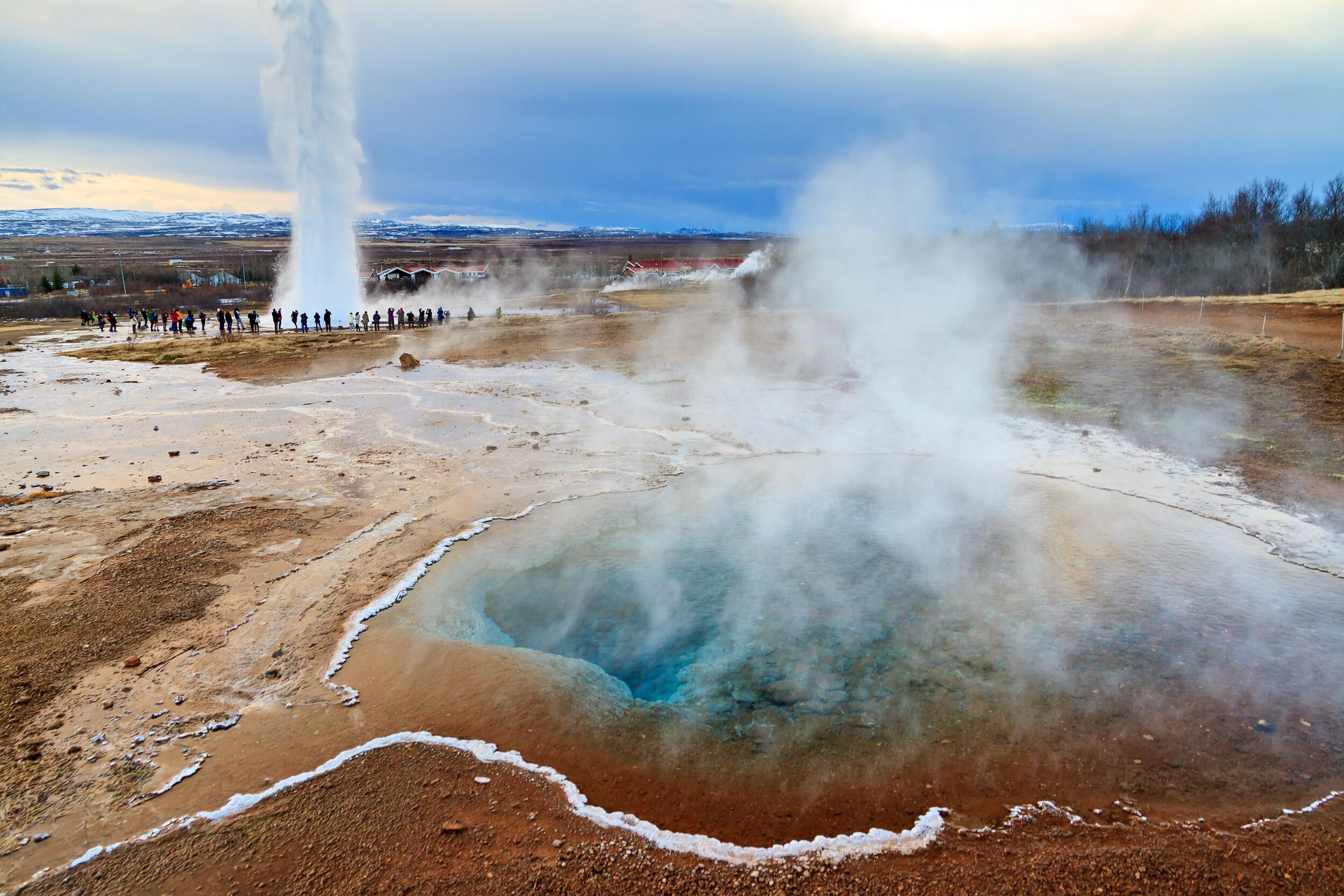 A vibrant geothermal hot spring with steaming turquoise water in the foreground and a powerful geyser erupting in the distance. A group of people stand nearby, observing this natural wonder, a highlight of a private Golden Circle tour.