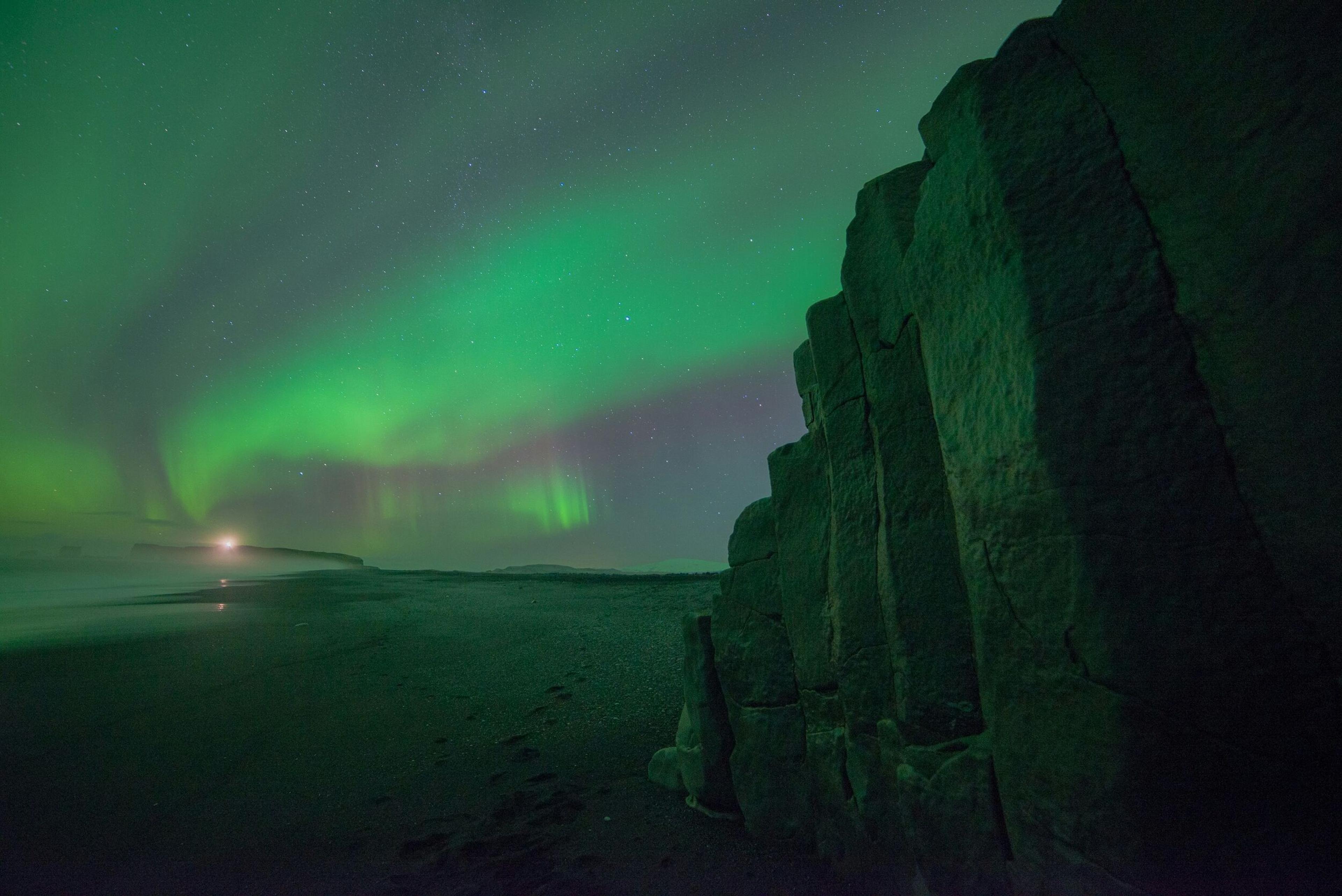 The Northern Lights illuminate the sky over a rocky shoreline, with the glow of the auroras casting a greenish hue on the landscape.