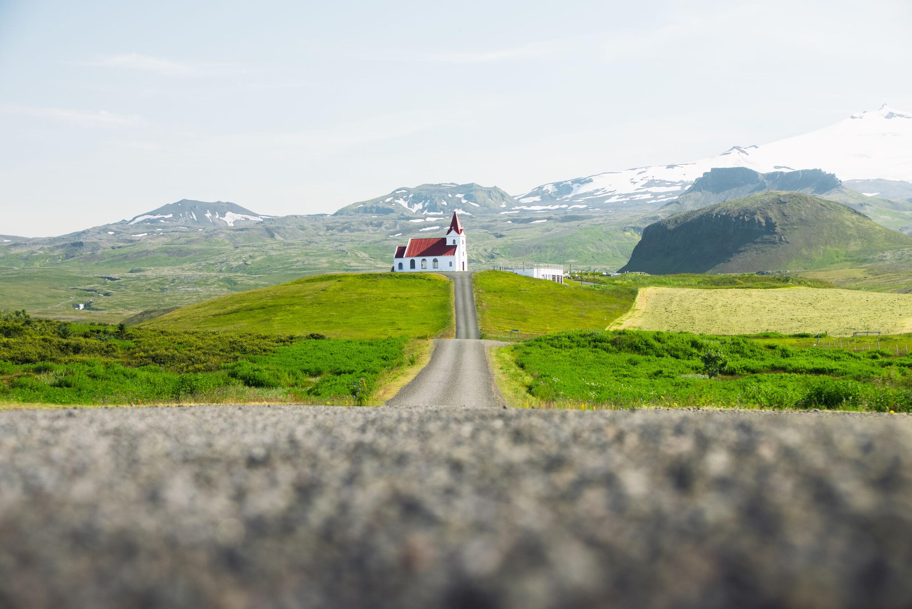 A road leading to a white church with a red roof on a green hill, with snowy mountains in the background.