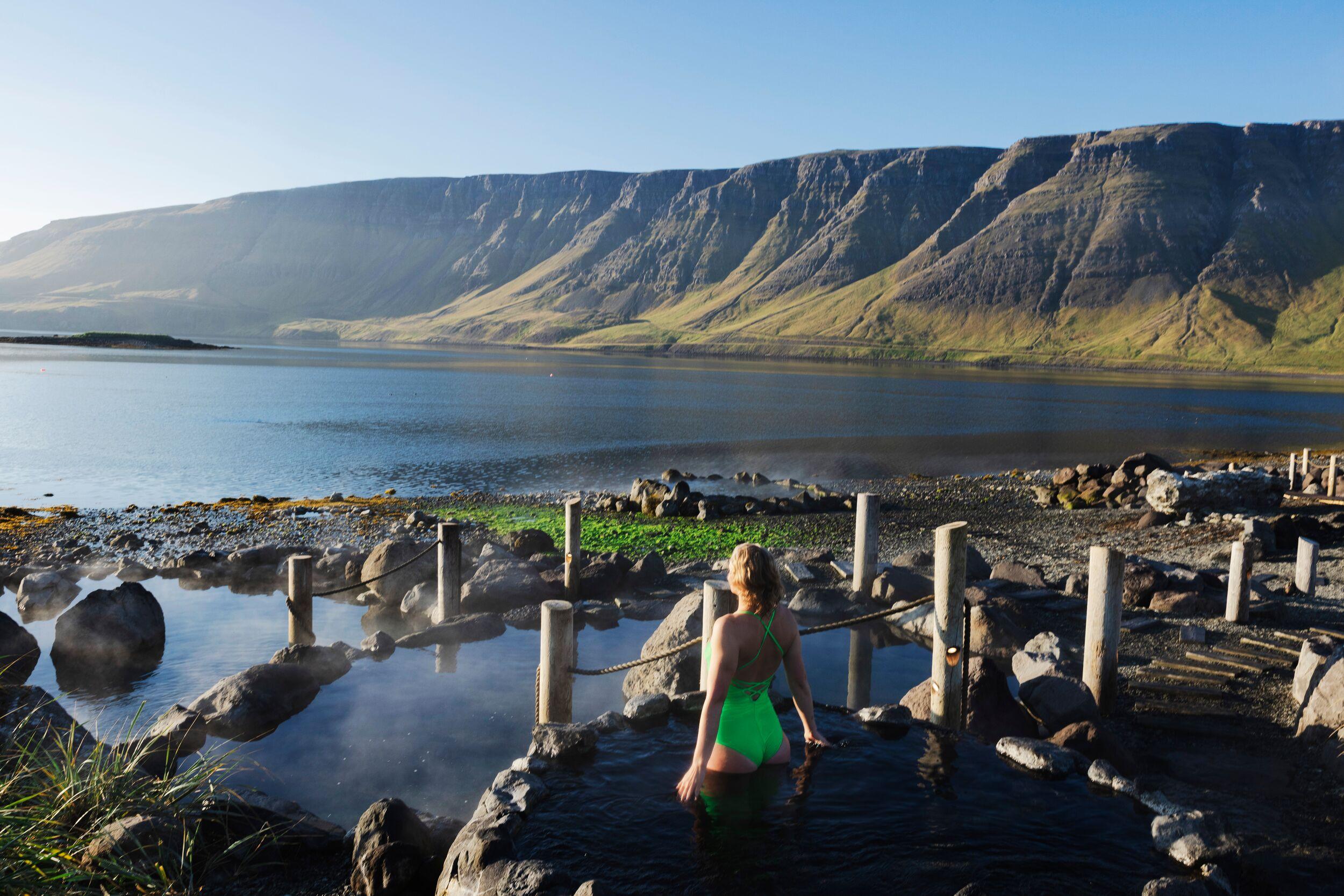 Woman in a green swimsuit in a steaming natural hot spring by a fjord with mountains.