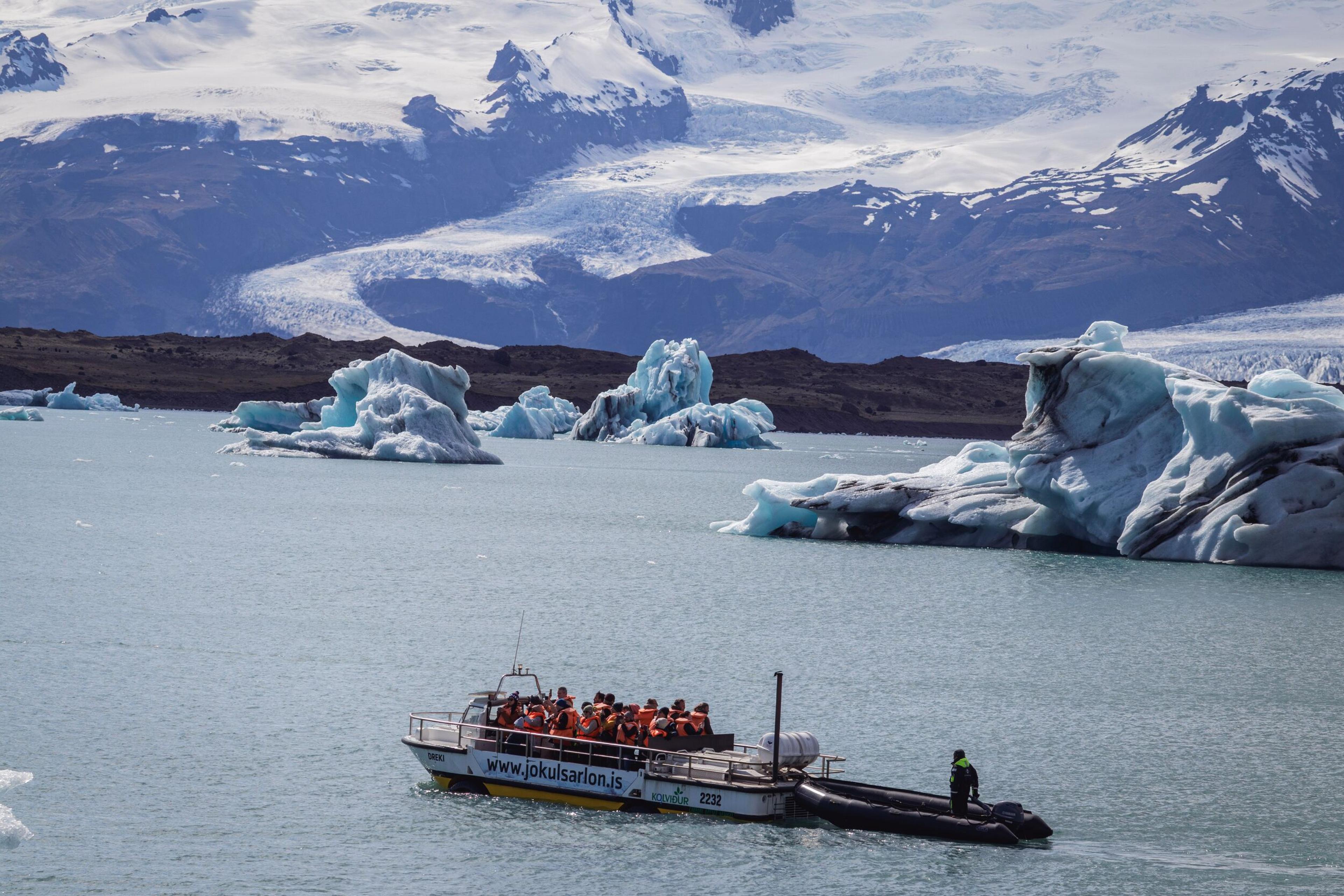 A tour boat full of passengers navigates a glacial lagoon filled with icebergs, backed by snow-covered mountains and a glacier.