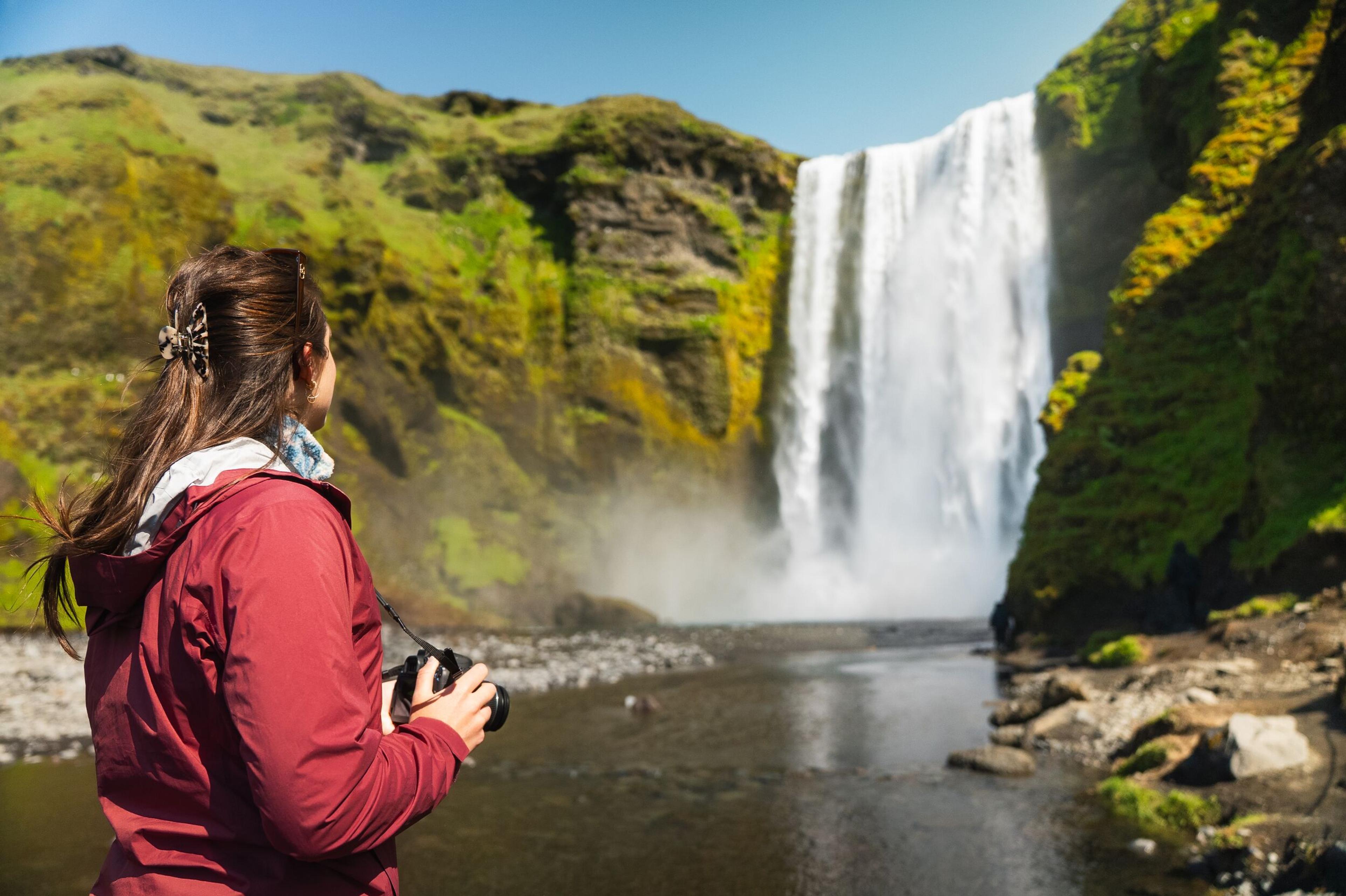 Woman in a red jacket holding a camera, admiring a large waterfall surrounded by green hills.