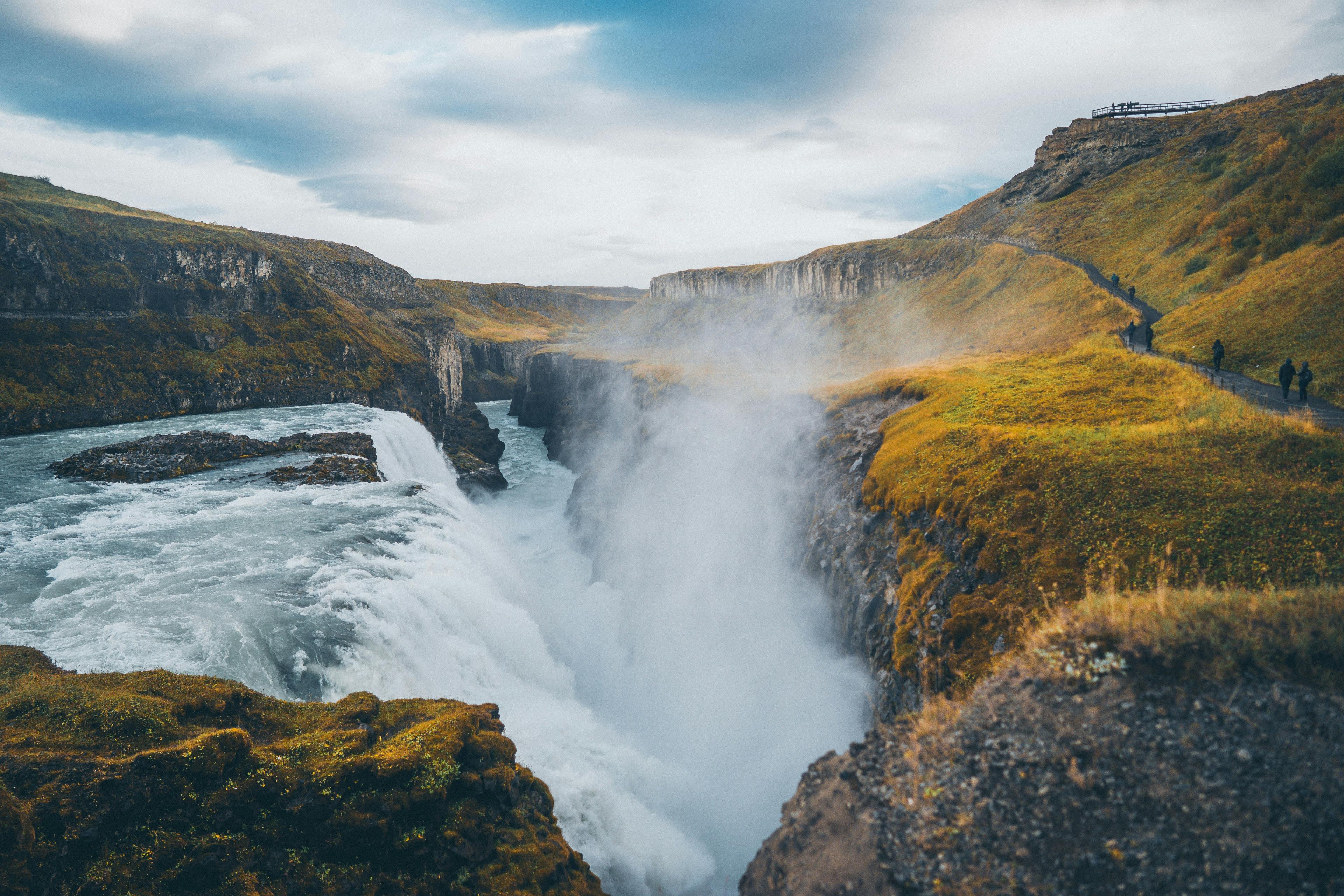 A powerful waterfall plunging into a canyon, creating a thick mist, with observers visible on a viewing platform along the verdant cliffs, a sight often sought after in the Golden Circle of Iceland.