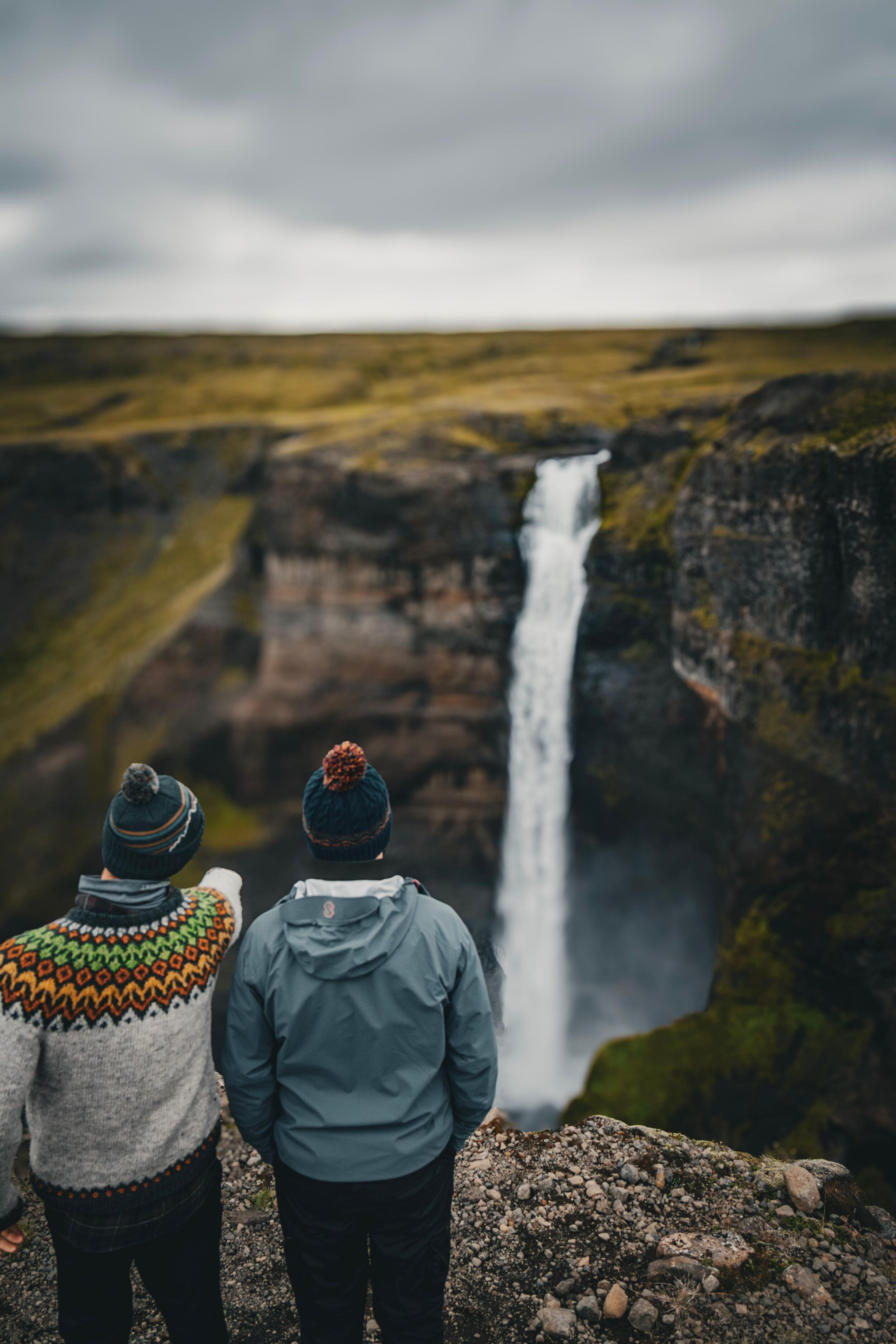 Two people in hats stand on a rocky overlook, one pointing at a tall waterfall plunging into a rugged canyon.