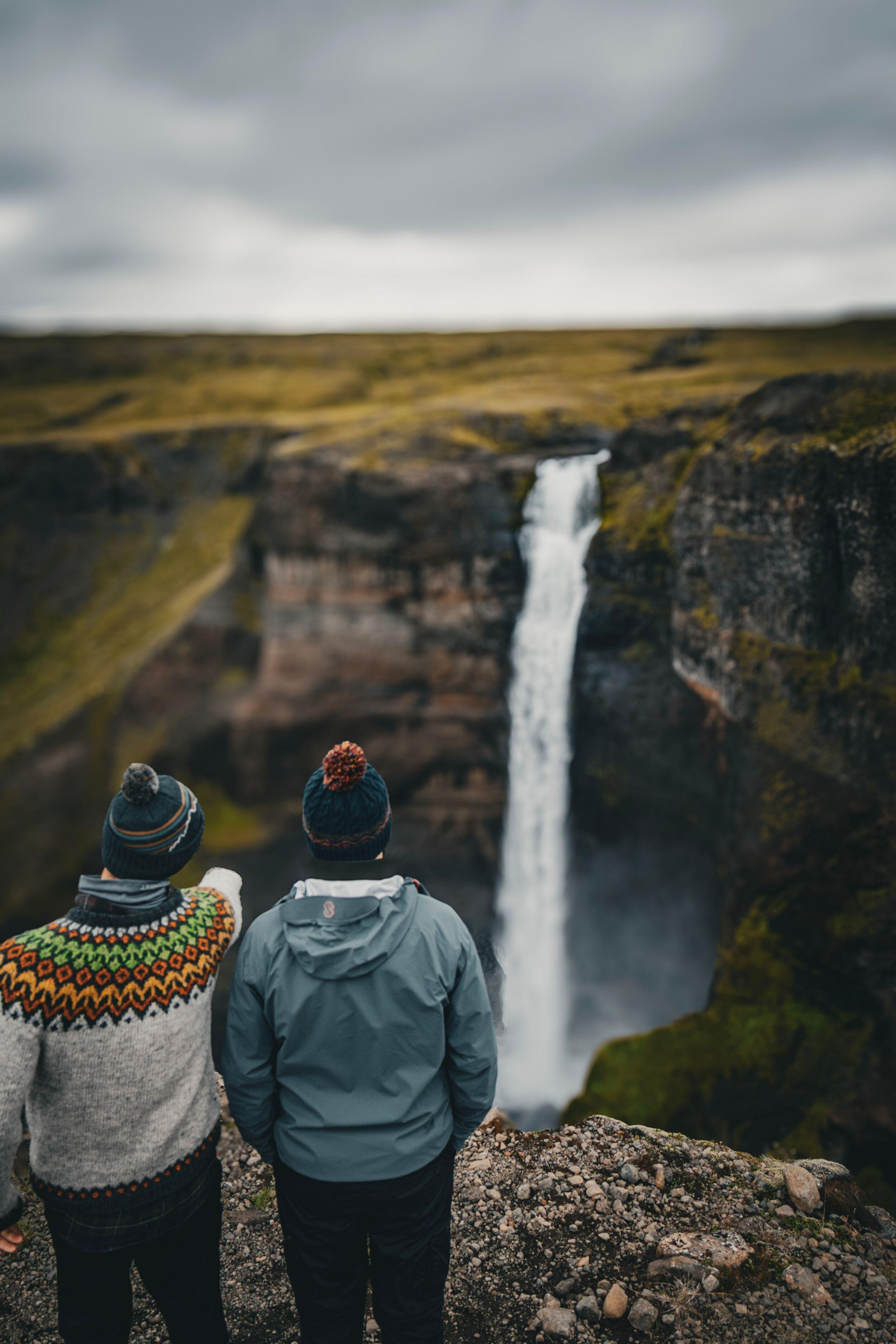 Two people in hats stand on a rocky overlook, one pointing at a tall waterfall plunging into a rugged canyon.