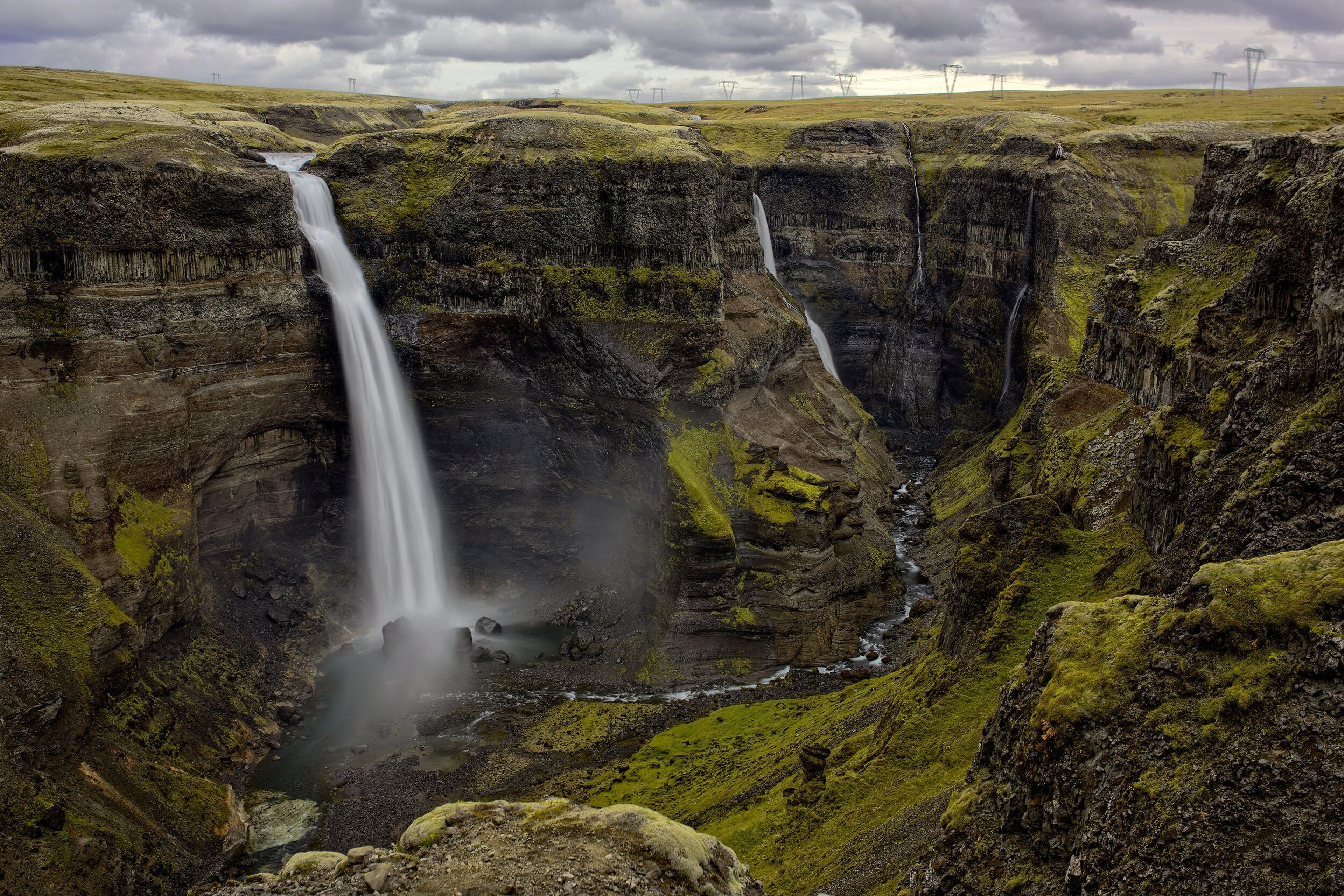 Two waterfalls plunge into a deep, mossy canyon with rocky walls under a cloudy sky.