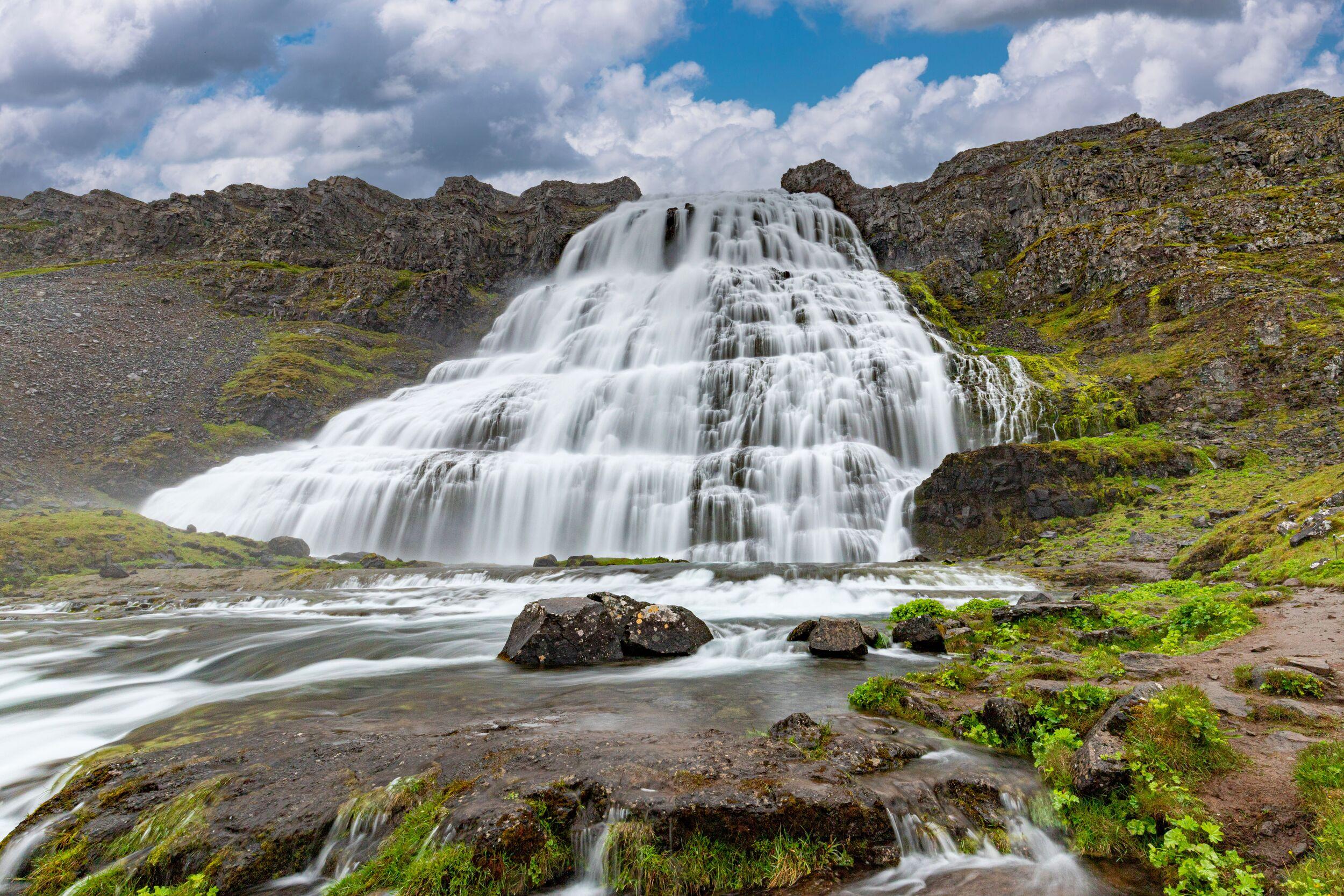 A multi-tiered waterfall cascades down a rocky, green cliff with long-exposure water, under a cloudy sky.