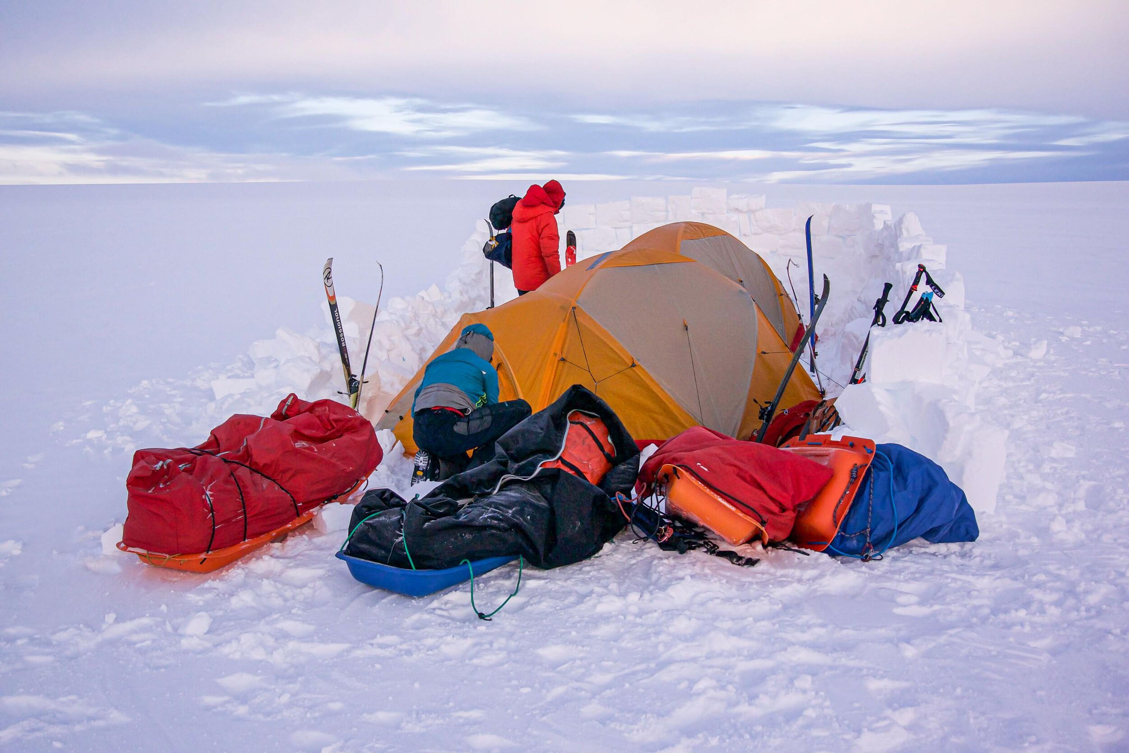 A campsite on a snowy expanse with a yellow tent surrounded by red and orange expedition gear and skis.