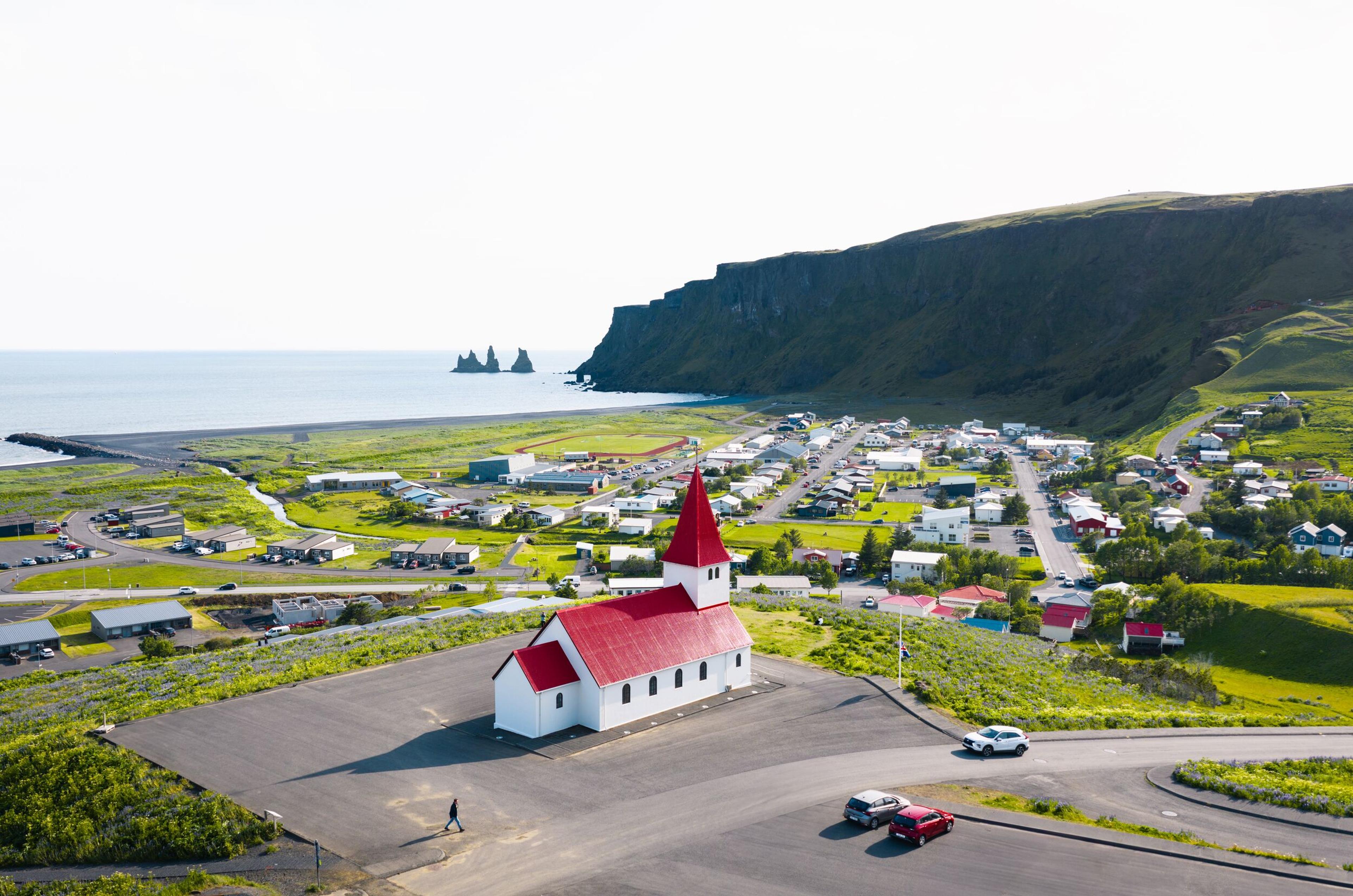 Aerial view of a white church with red roof overlooking a coastal village, black sand beach, sea stacks, and green cliffs.