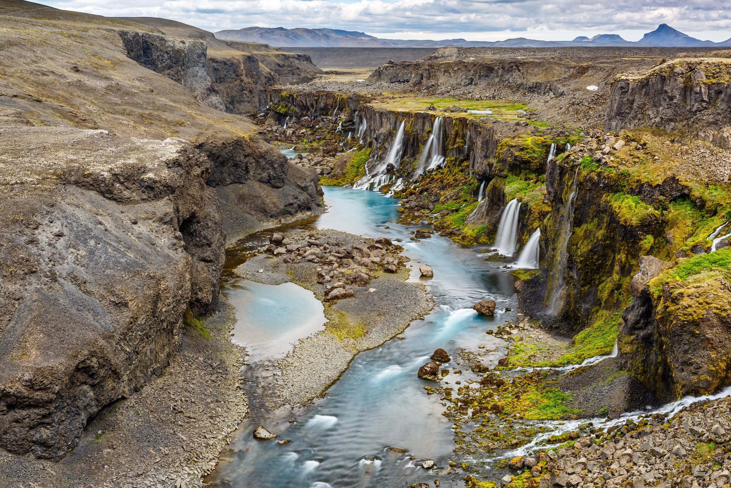 Overview of Sigöldugljúfur canyon in Iceland, featuring a winding river flanked by steep, moss-covered cliffs with numerous small waterfalls feeding into turquoise blue pools, set against a barren, volcanic landscape