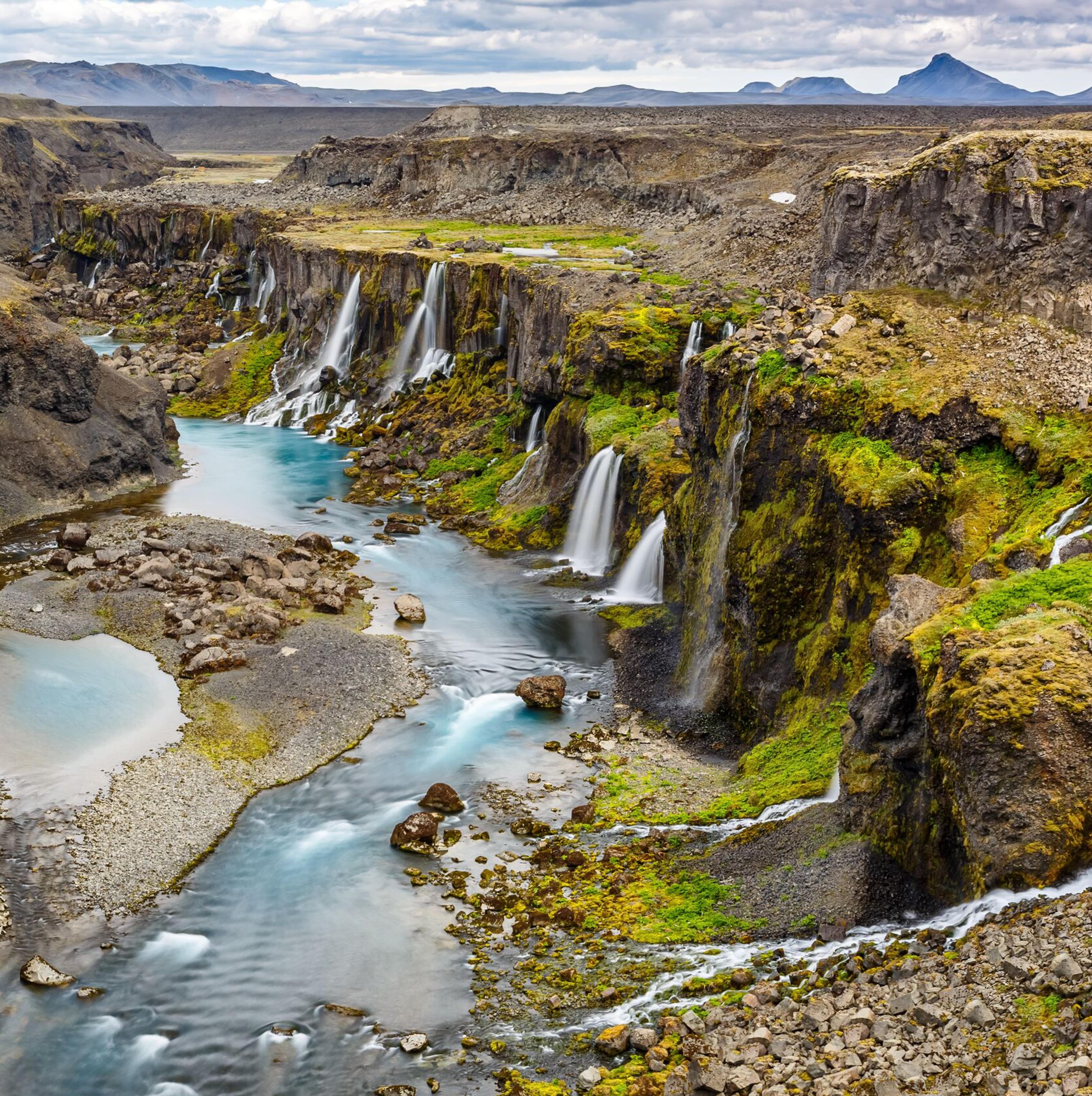 Overview of Sigöldugljúfur canyon in Iceland, featuring a winding river flanked by steep, moss-covered cliffs with numerous small waterfalls feeding into turquoise blue pools, set against a barren, volcanic landscape