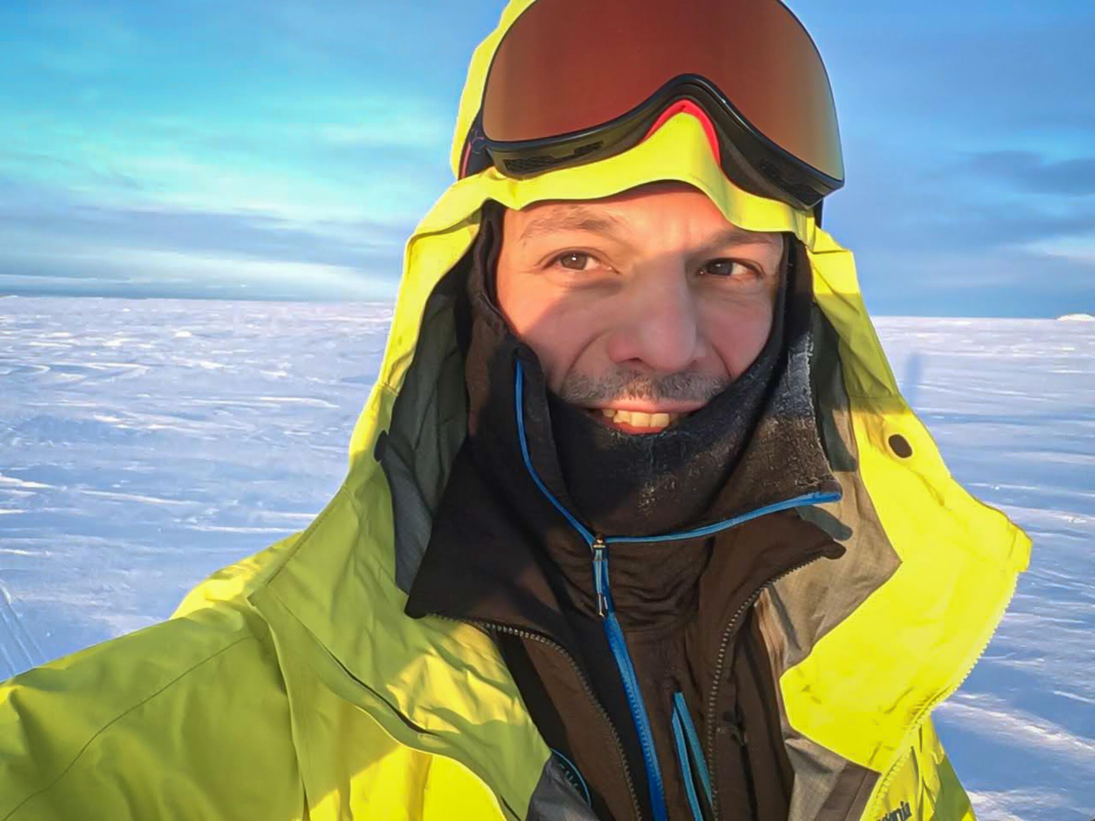 A selfie of a person in cold-weather gear with a hood and goggles, with a vast expanse of snow and ice stretching out behind them under a bright sky.