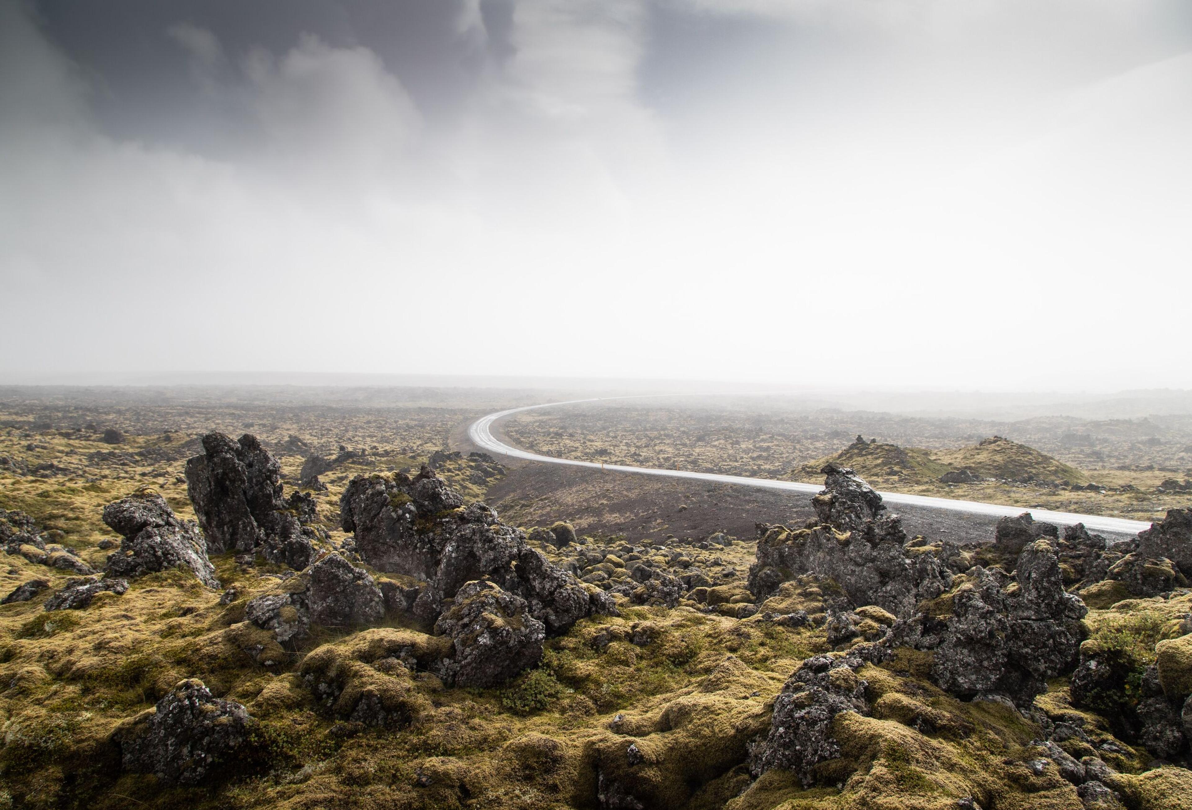 A curving road cuts through a stark, rocky landscape covered in moss, under a misty and overcast sky.
