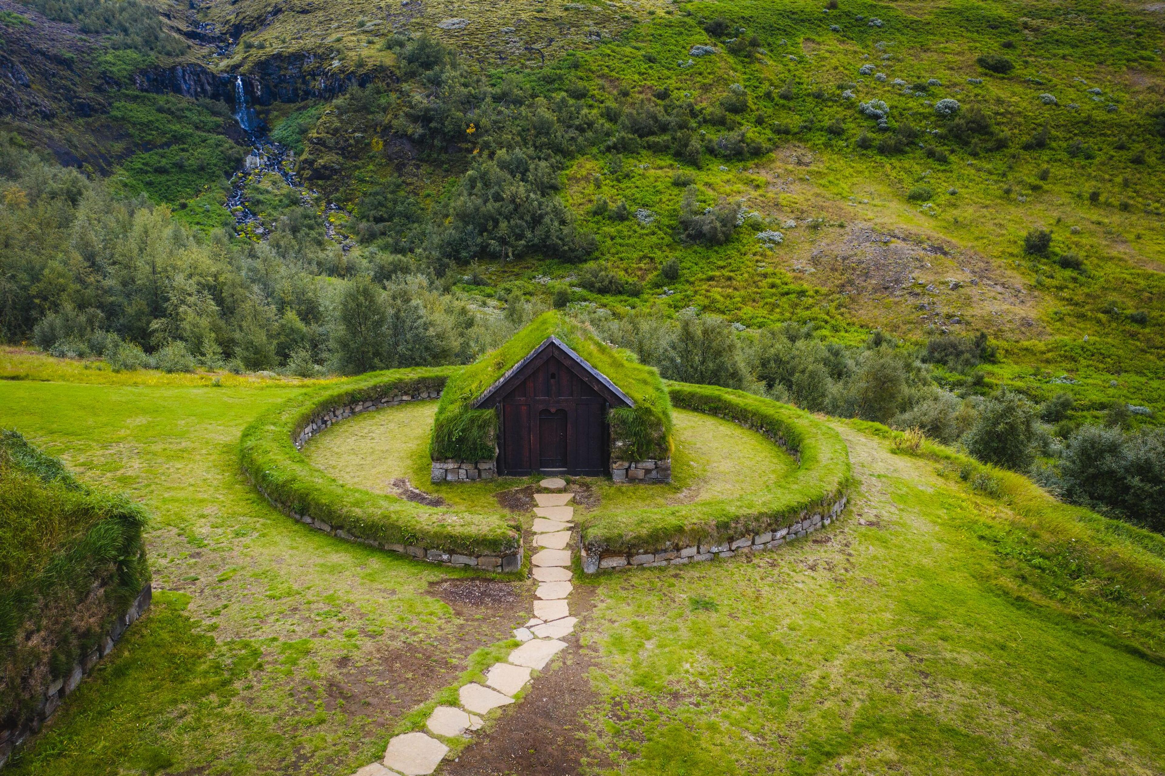 A grass-roofed wooden building with a stone path, encircled by a low grass wall on a green hill with a waterfall.