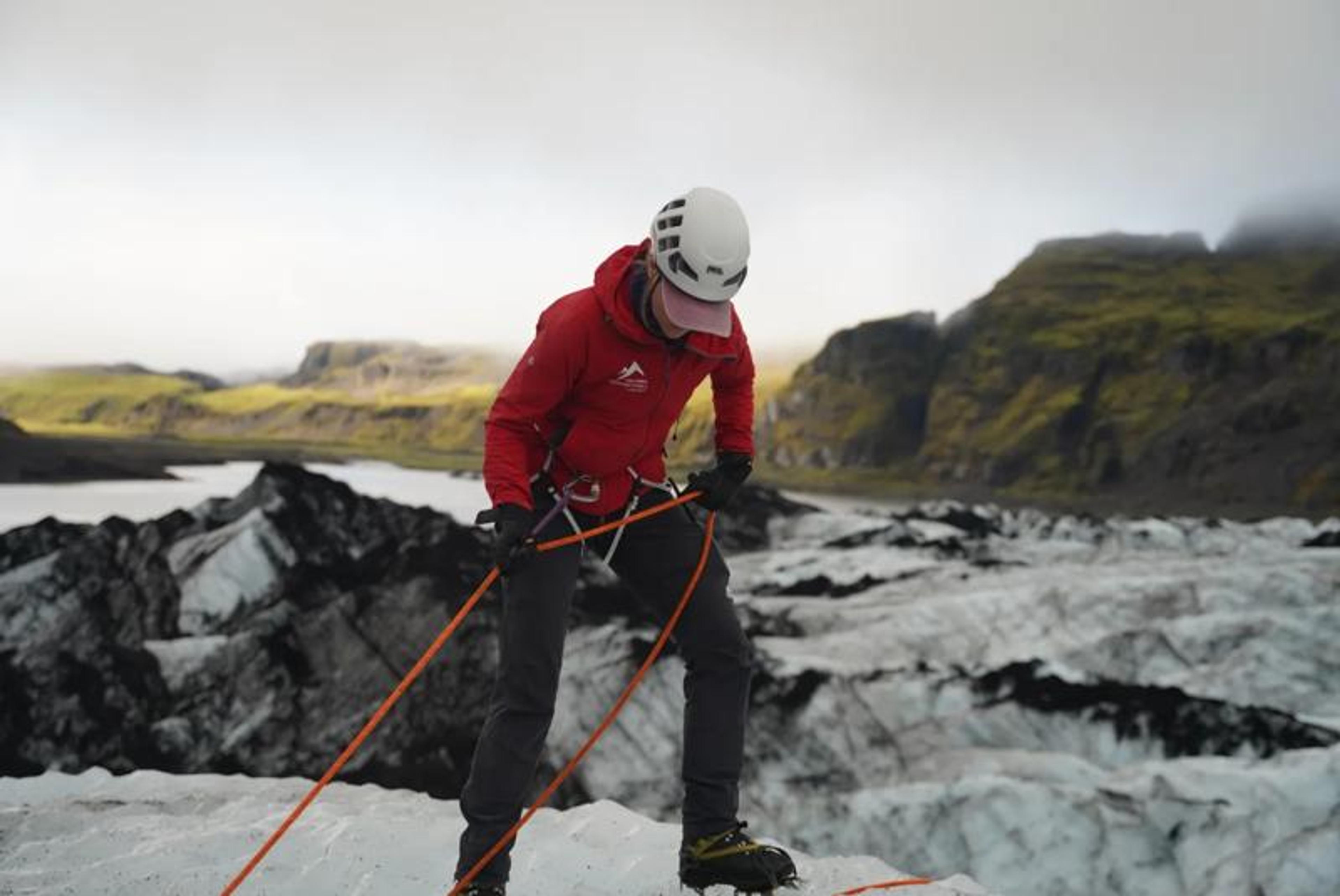 A person in a red jacket and helmet stands on a glacier, wearing a climbing harness and holding ropes, with dark mountains and a lake in the background.
