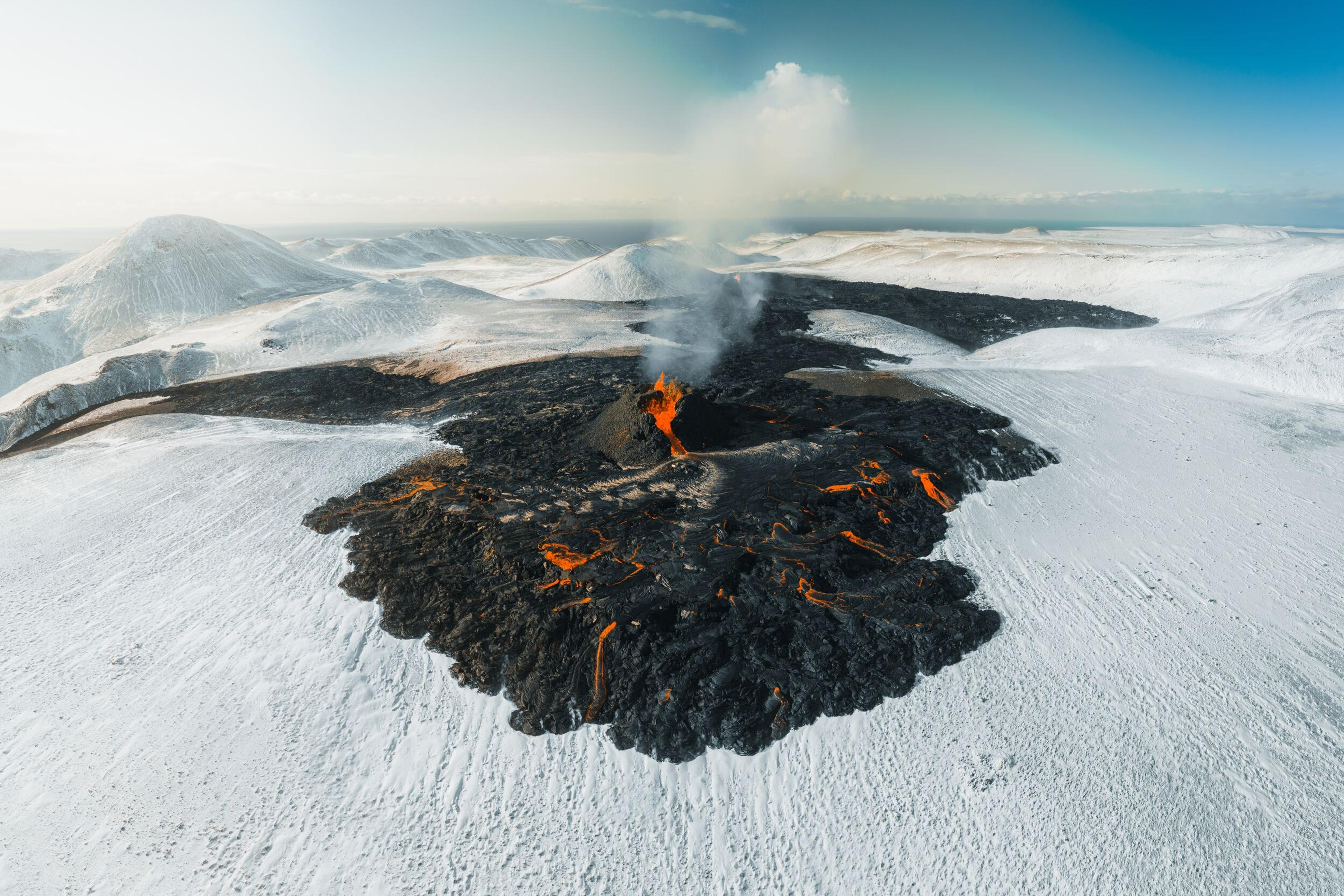 Icelandic volcano with orange lava erupting, surrounded by snow-covered ground, under a clear sky.