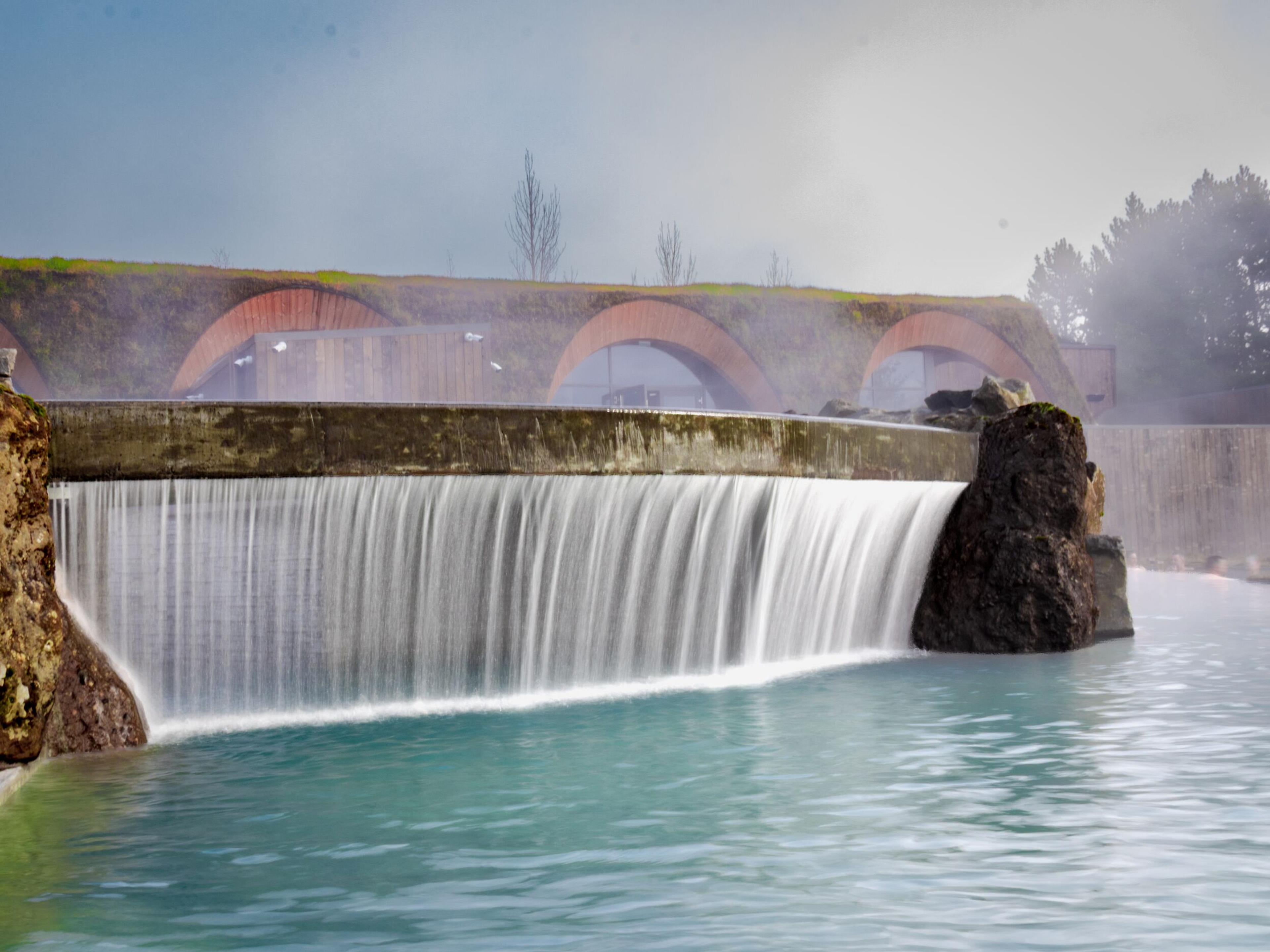 Waterfall flowing into a blue pool, with arch-shaped buildings integrated into a grassy hillside.