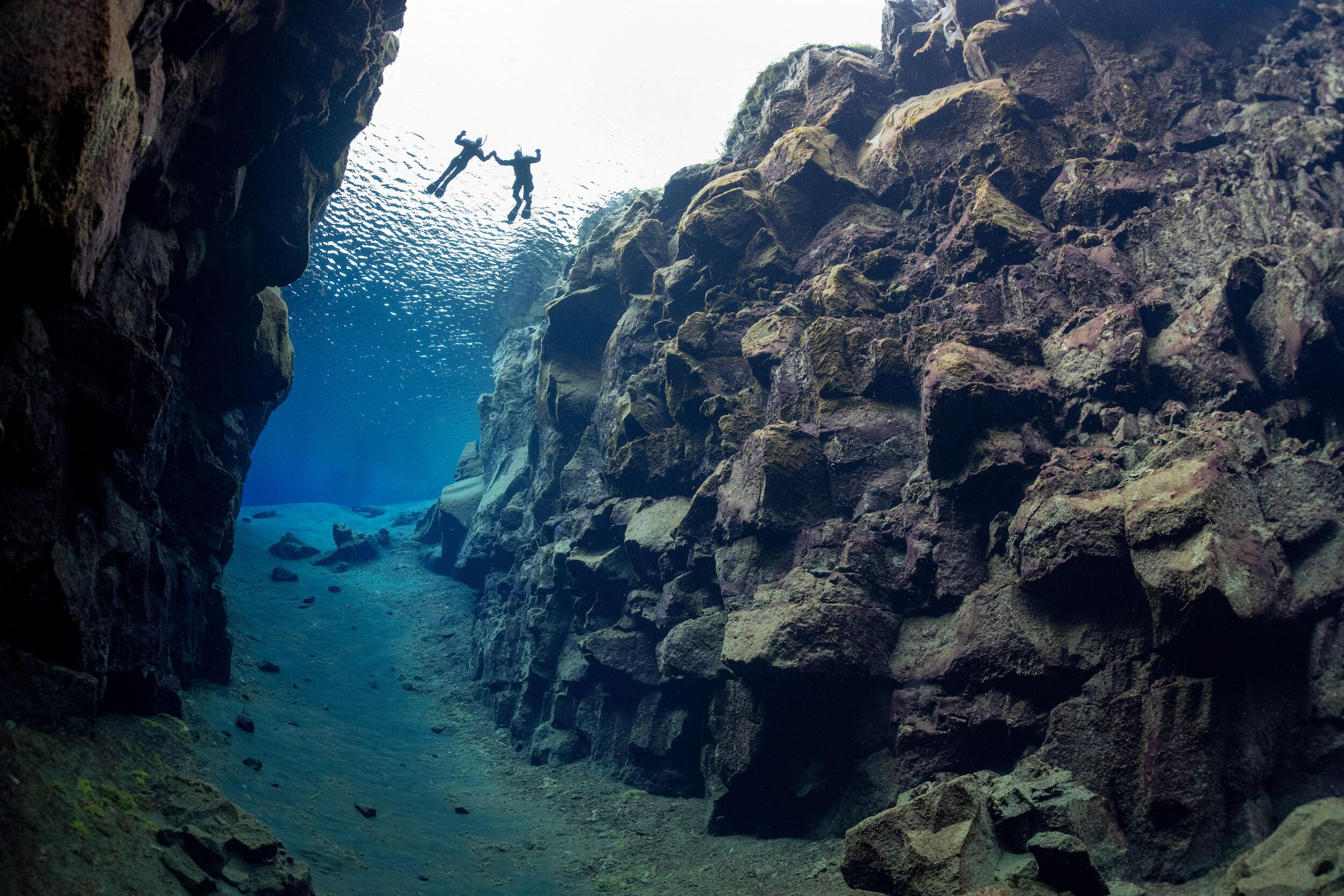 Two divers float in crystal-clear blue water within a narrow underwater rift between rugged rock walls.