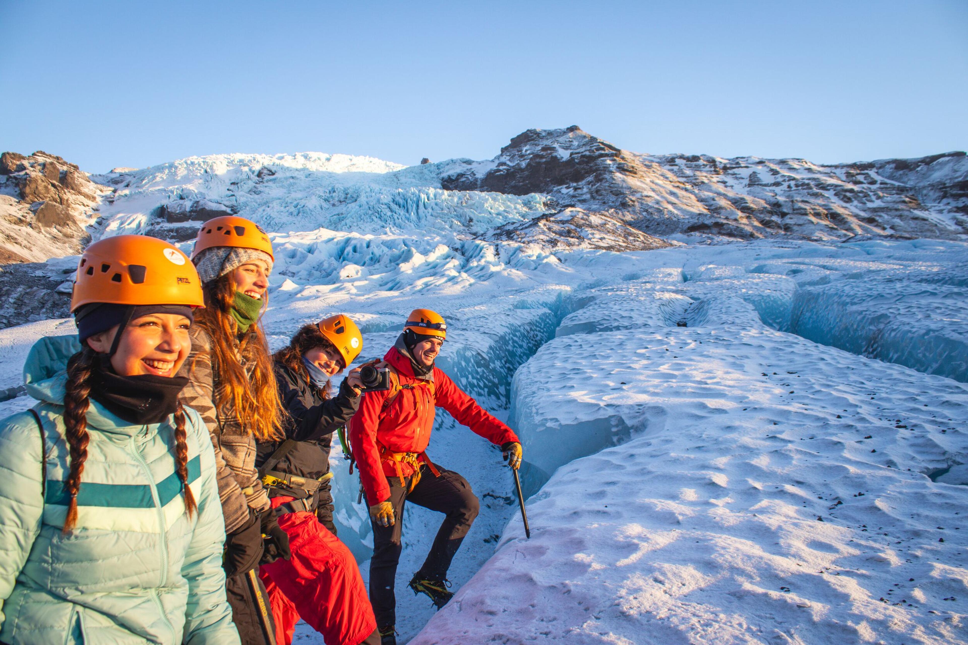 Four people in helmets on a sunny glacier, with blue ice formations and snow-capped mountains.