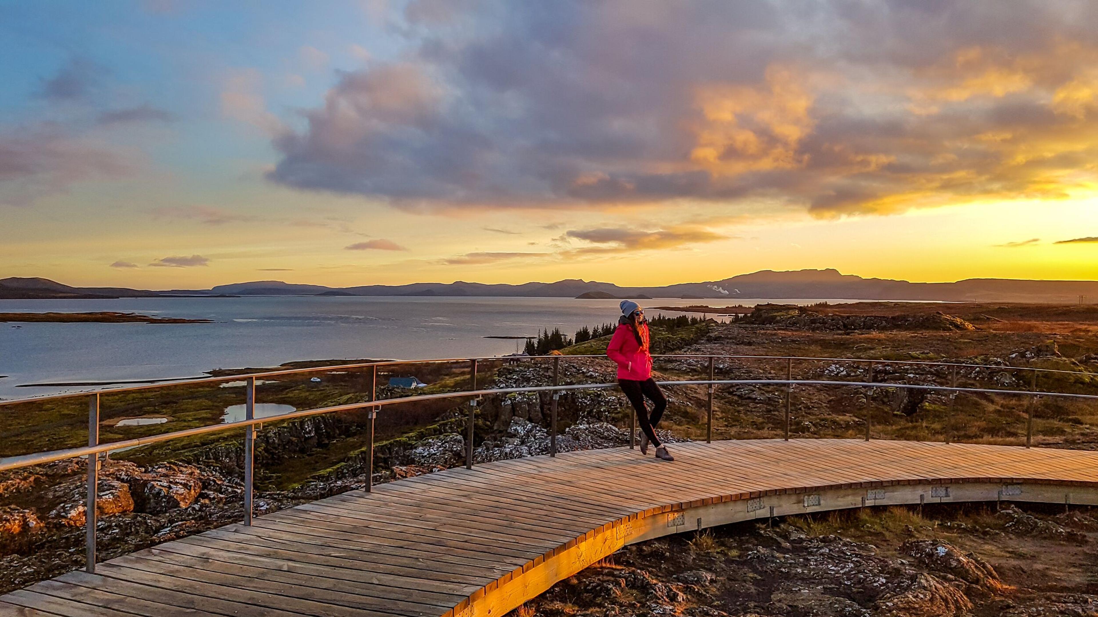 A person wearing a pink jacket and beanie stands on a curved wooden pathway overlooking a serene lake and rugged landscape at sunset. The sky is painted with vibrant shades of orange, yellow, and blue, reflecting the peaceful atmosphere.